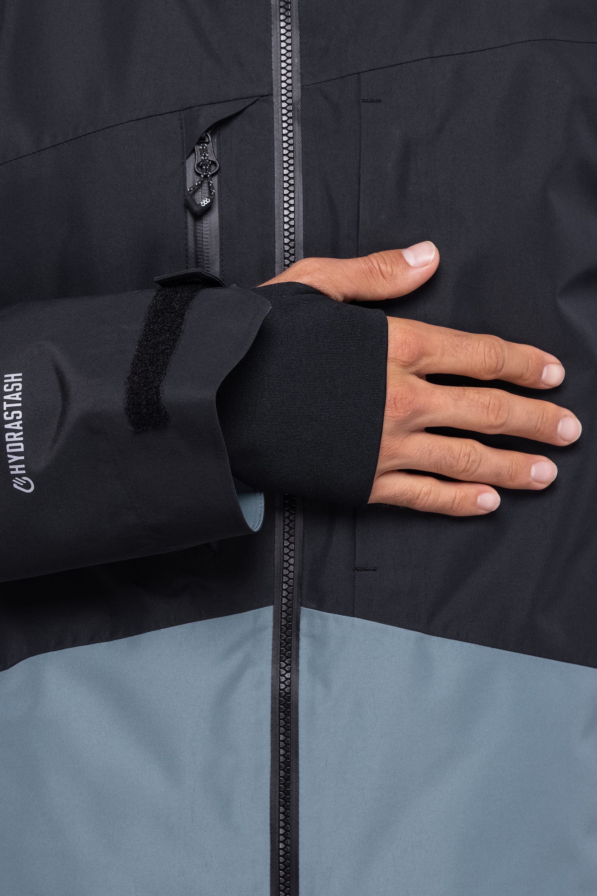 A close-up shows a person wearing the 686 Men's Hydrastash Reserve Insulated Jacket in black and gray, with their right hand on their chest. The sleeve, featuring a thumbhole, displays the HYDRASTASH logo.