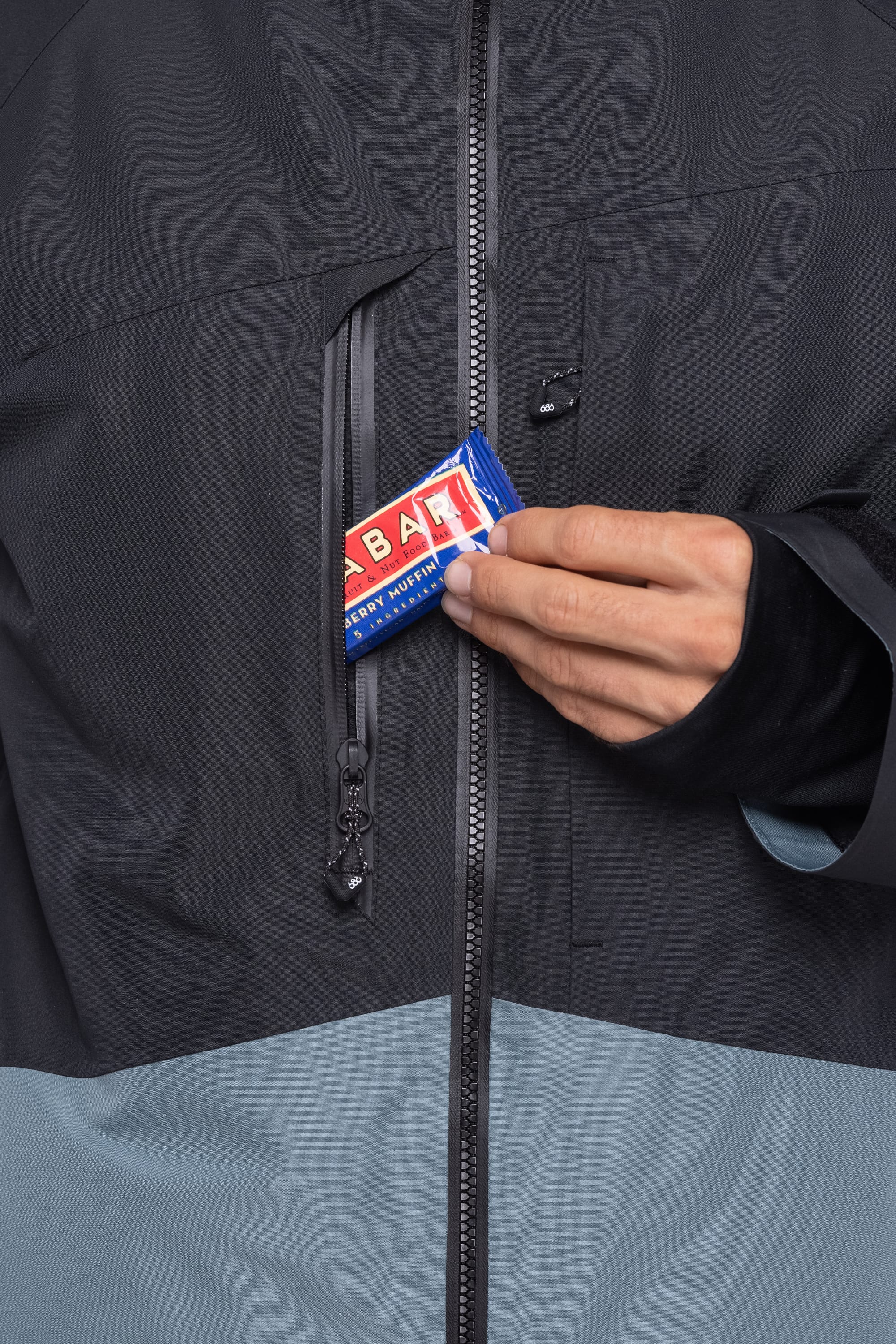 A person is placing a blue-wrapped Clif Bar into the chest pocket of a 686 Men's Hydrastash Reserve Insulated Jacket, with only their hand and part of the bar visible.