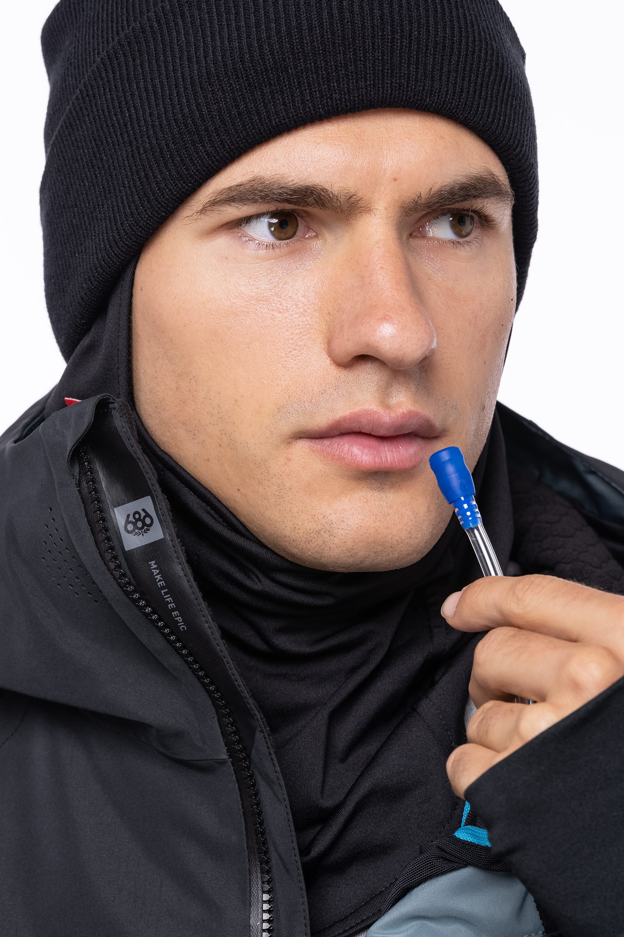 A man in the 686 Men's Hydrastash Reserve Insulated Jacket wears a black beanie and holds the blue tip of a hydration tube near his mouth, looking sideways with a focused expression.