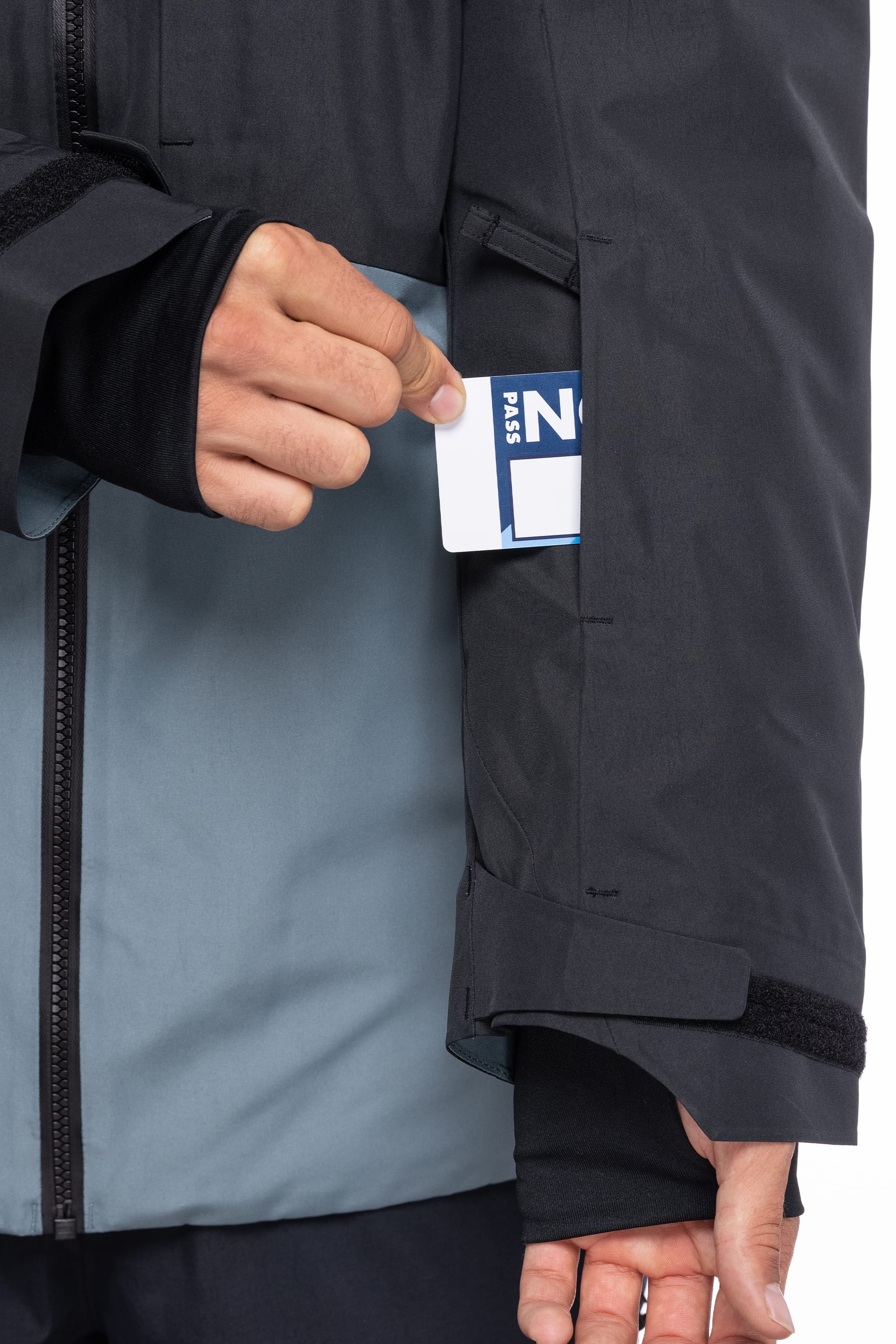 A person slips a ski pass into the zippered sleeve pocket of the 686 Men's Hydrastash Reserve Insulated Jacket in black and gray, with their hand partly covered by the thumbhole cuff.