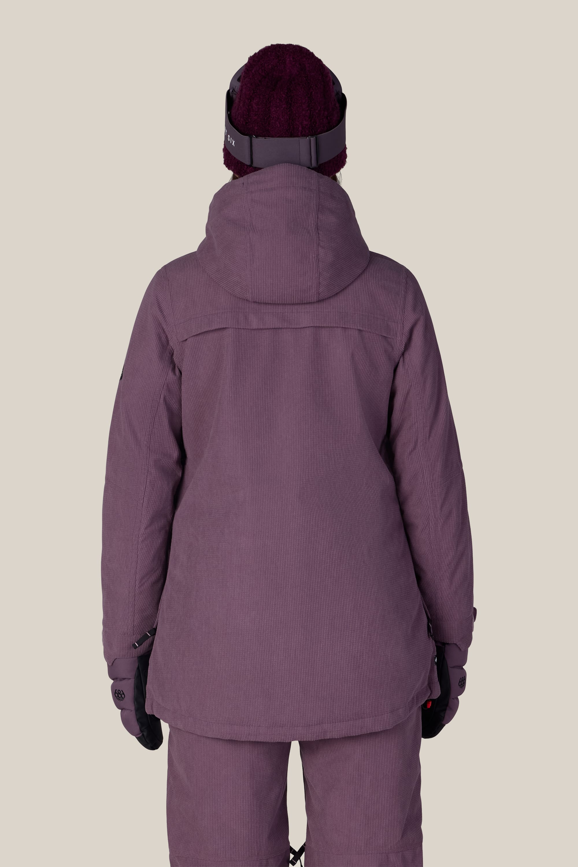 A woman is shown from the back against a plain beige background wearing the 686 Women’s Spirit Insulated Jacket in purple, paired with matching pants, gloves, and a knit hat.