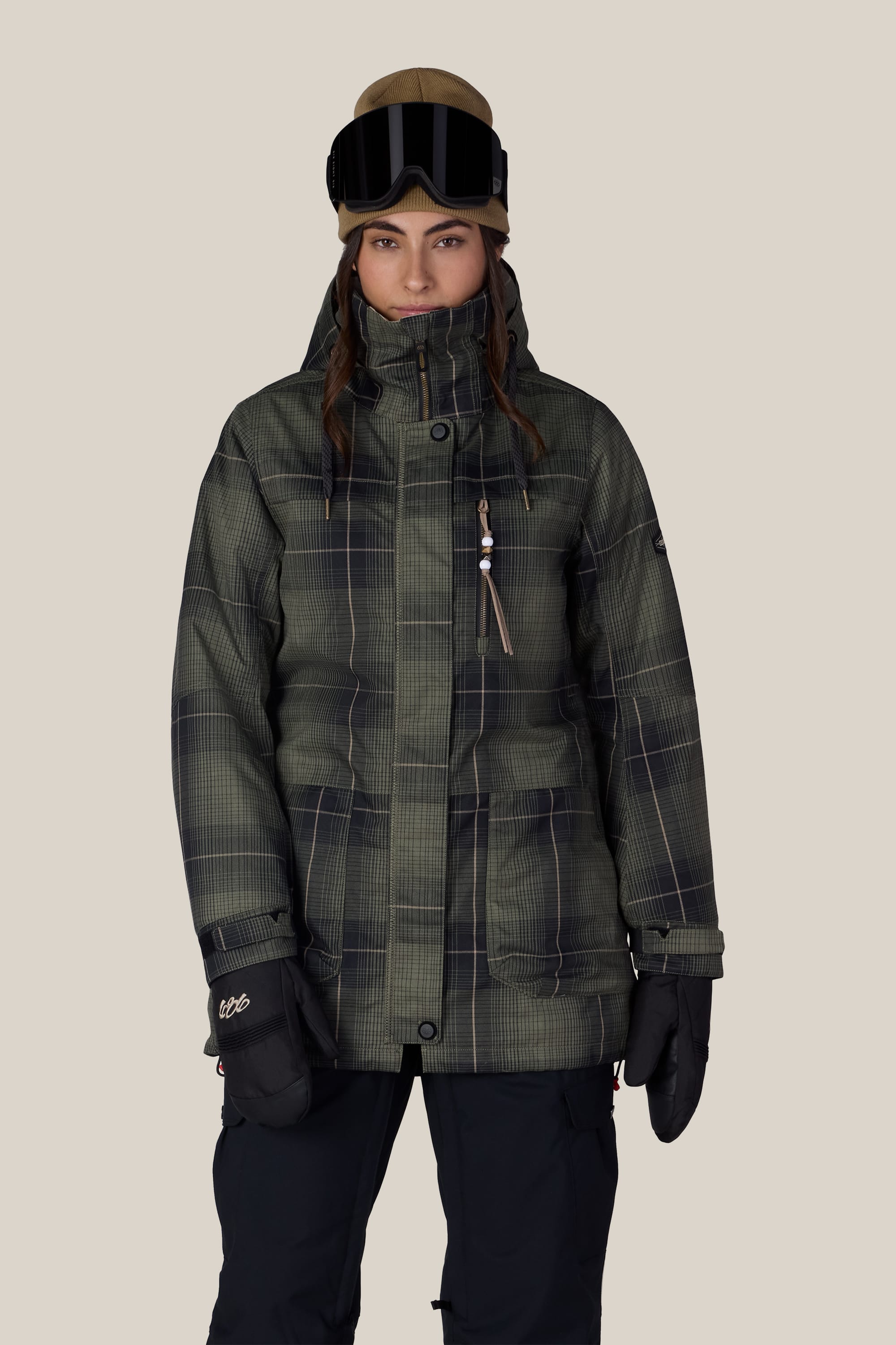 A person wearing the 686 Women’s Spirit Insulated Jacket, black snow pants, ski gloves, and ski goggles on their head stands against a plain beige background.