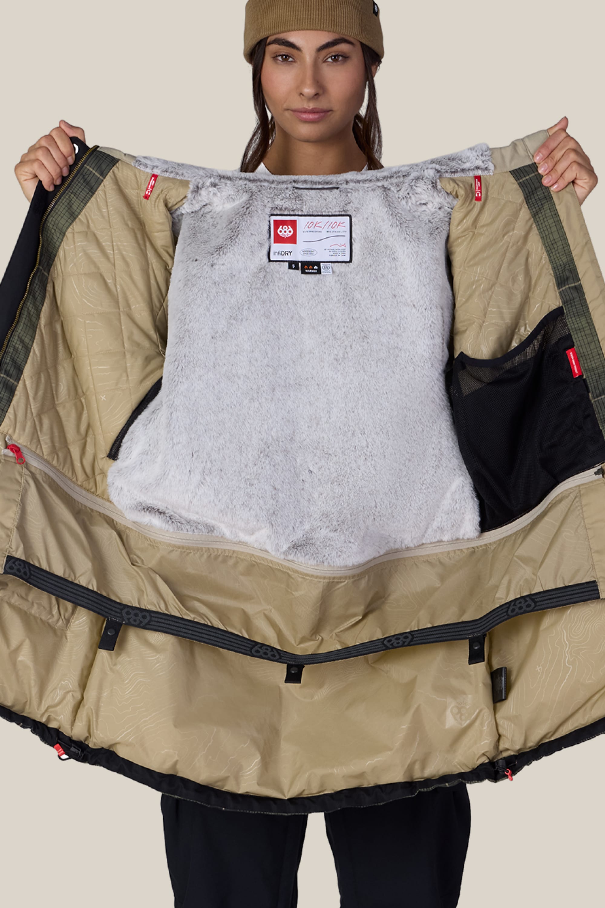 A person wearing a tan beanie holds open the 686 Women’s Spirit Insulated Jacket, showcasing its soft gray inner lining, multiple interior pockets, and a 686 brand label near the neckline.