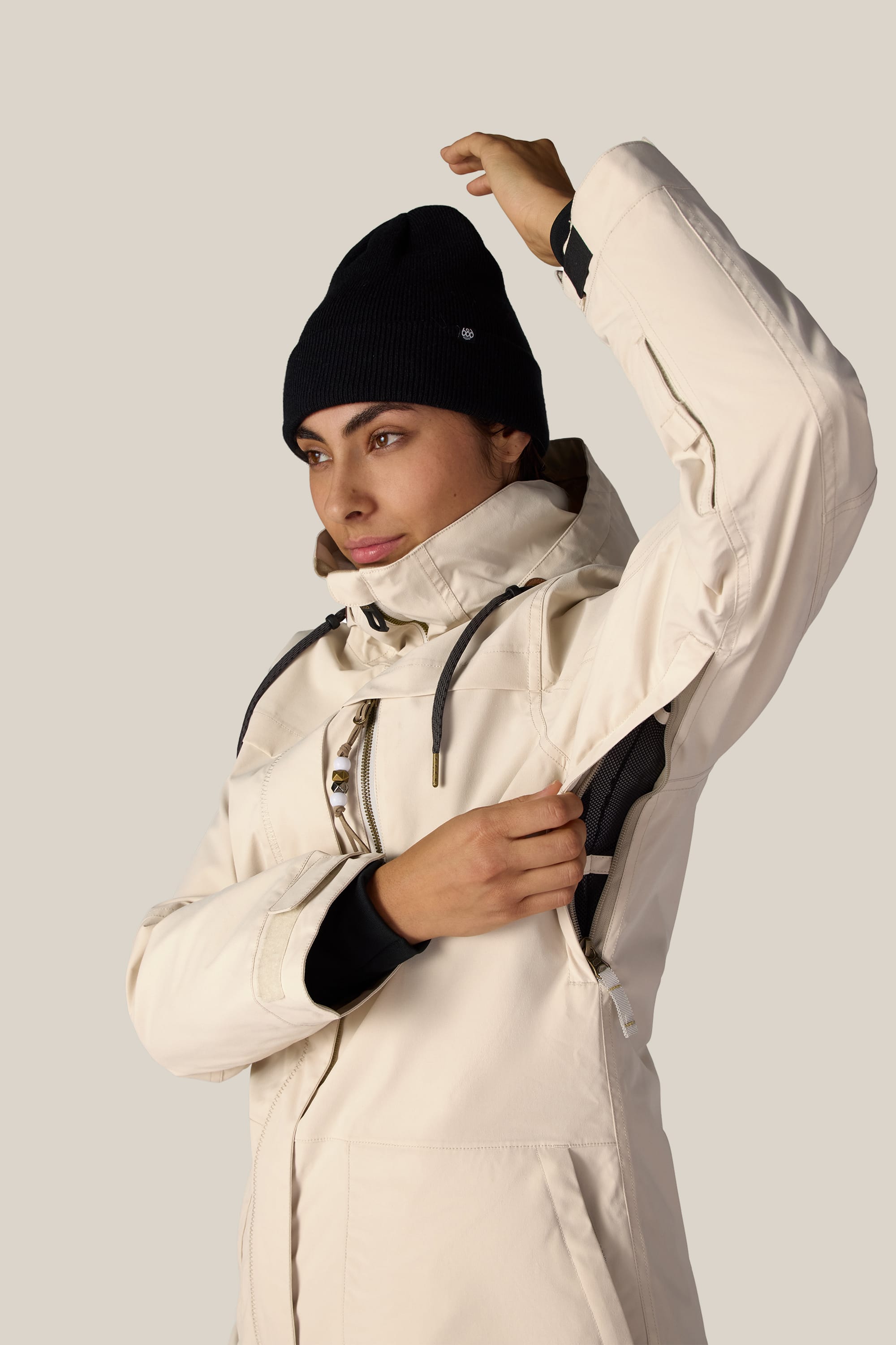 A person wearing the 686 Women’s Spirit Insulated Jacket in cream and a black beanie lifts their right arm, using their left hand to unzip the underarm vent. The background is plain and light-colored.