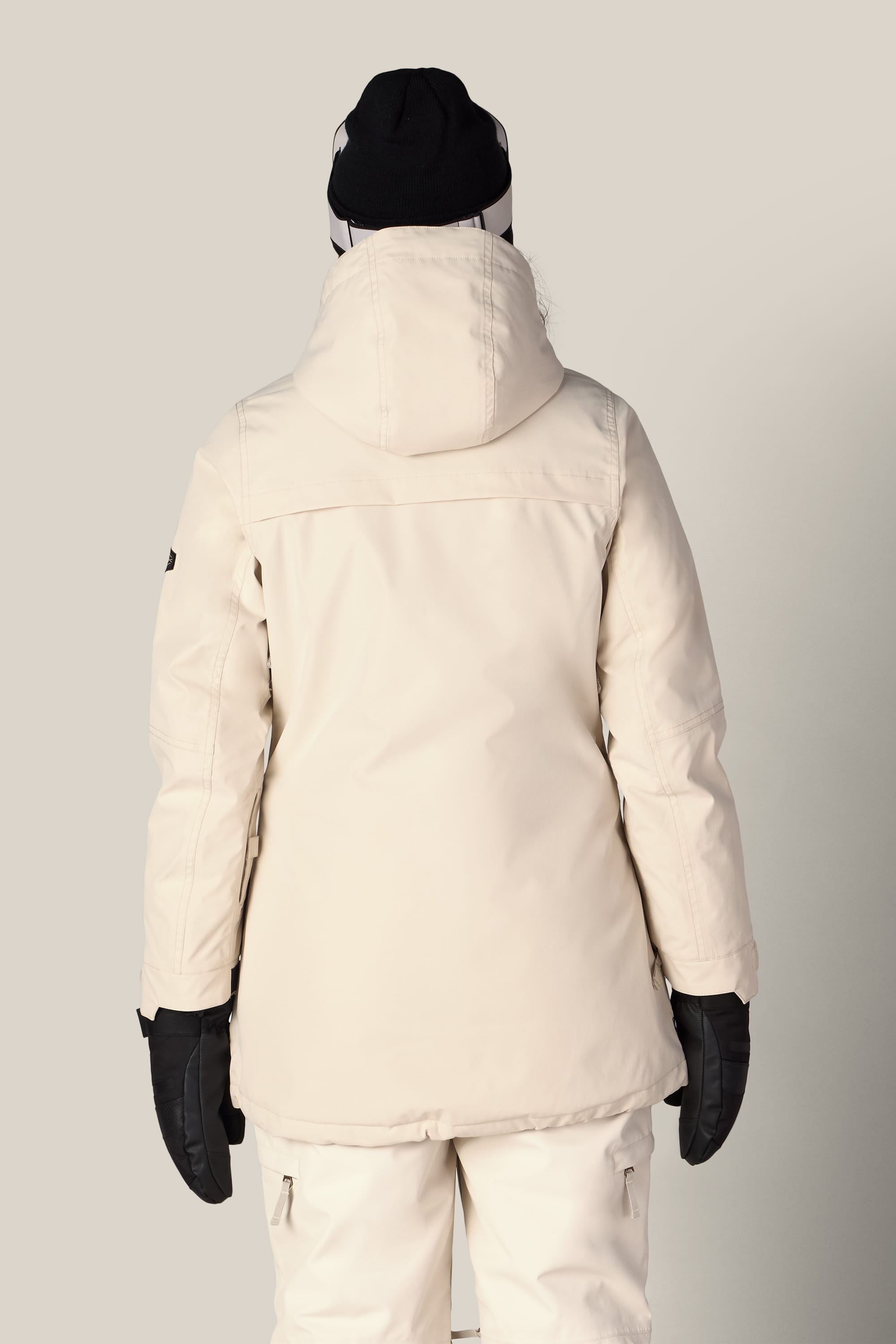 A person is facing away, wearing the 686 Women’s Spirit Insulated Jacket in a cream color, paired with matching pants, black gloves, and a black beanie. The background is plain light grey.