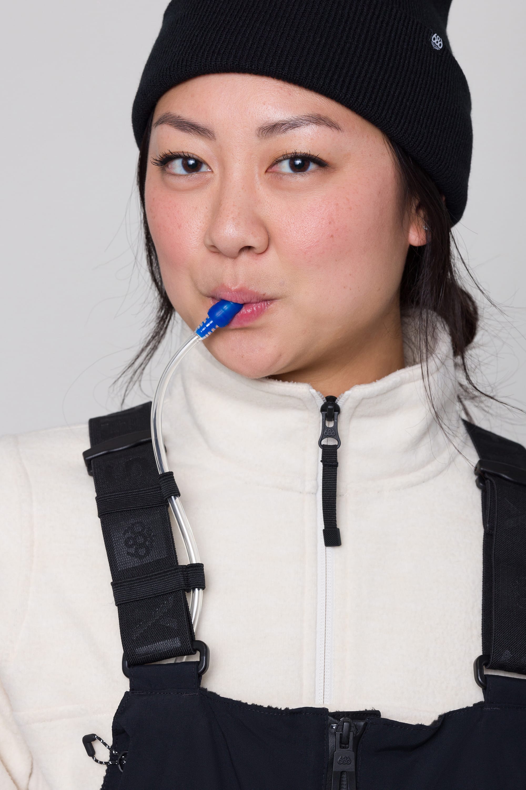 A woman in a black beanie and white zip-up jacket sips from a 686 Womens Complete Bladder System hydration tube attached to her black overalls, looking into the camera with a slight smile.
