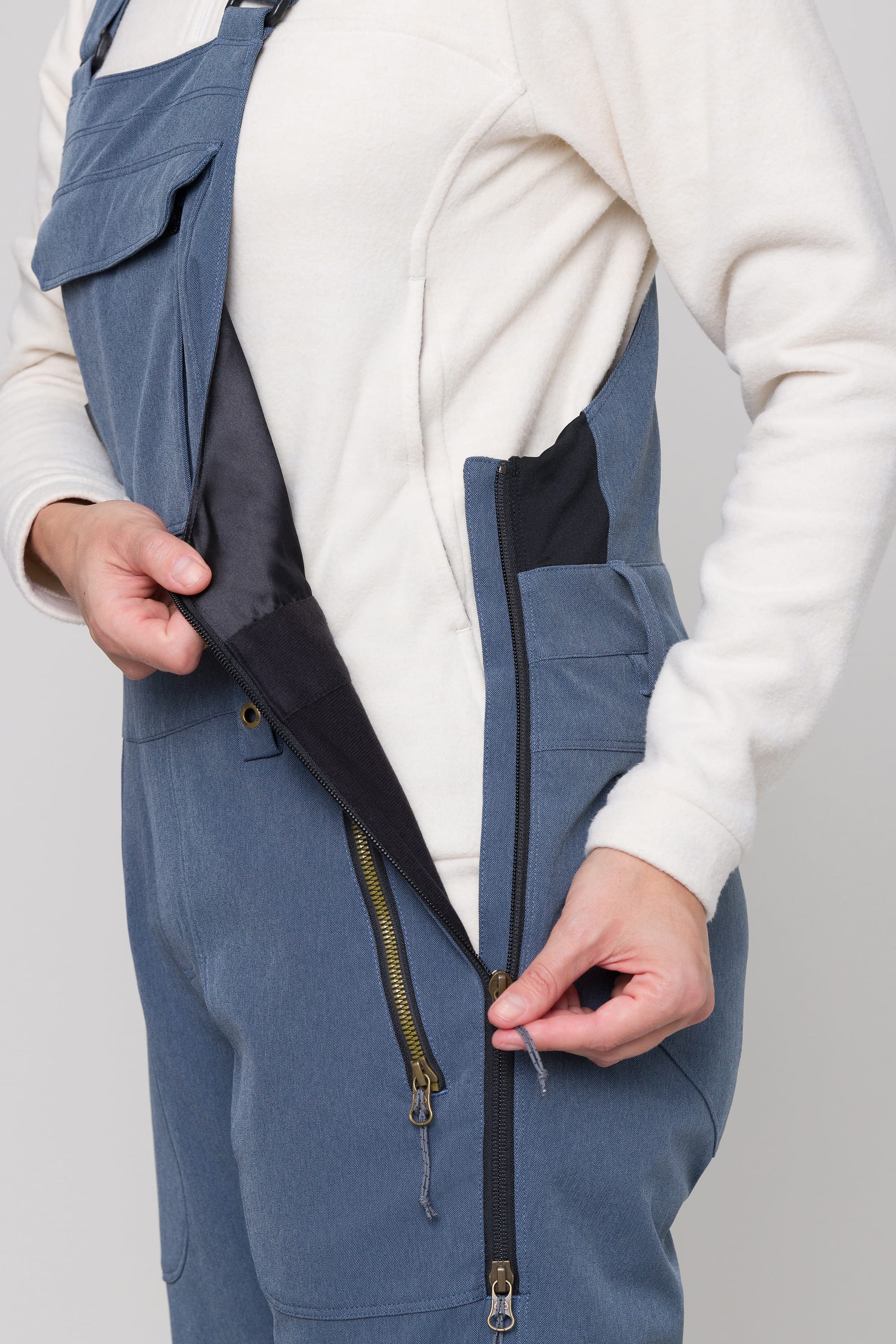 A person, face not visible, in a white long-sleeve shirt demonstrates unzipping the side of the 686 Womens Black Magic Insulated Bib by 686, revealing the inner lining and zipper mechanism.