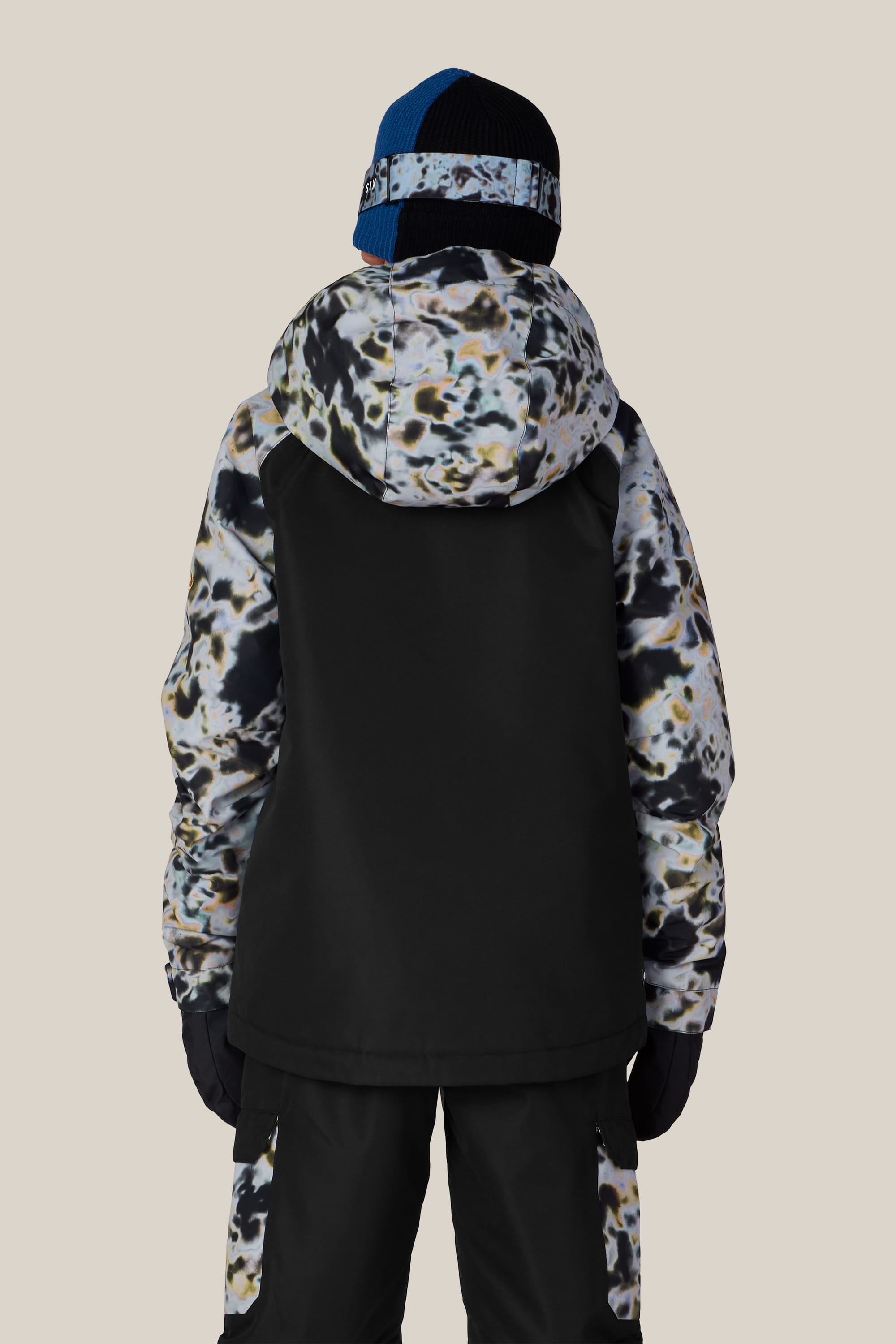 A person is wearing the 686 Boys’ Geo™ Insulated Jacket by 686, featuring a black and multicolored camouflage design, paired with black pants, gloves, and a knit hat with a blue and black pattern. The individual is photographed from the back against a plain light background.