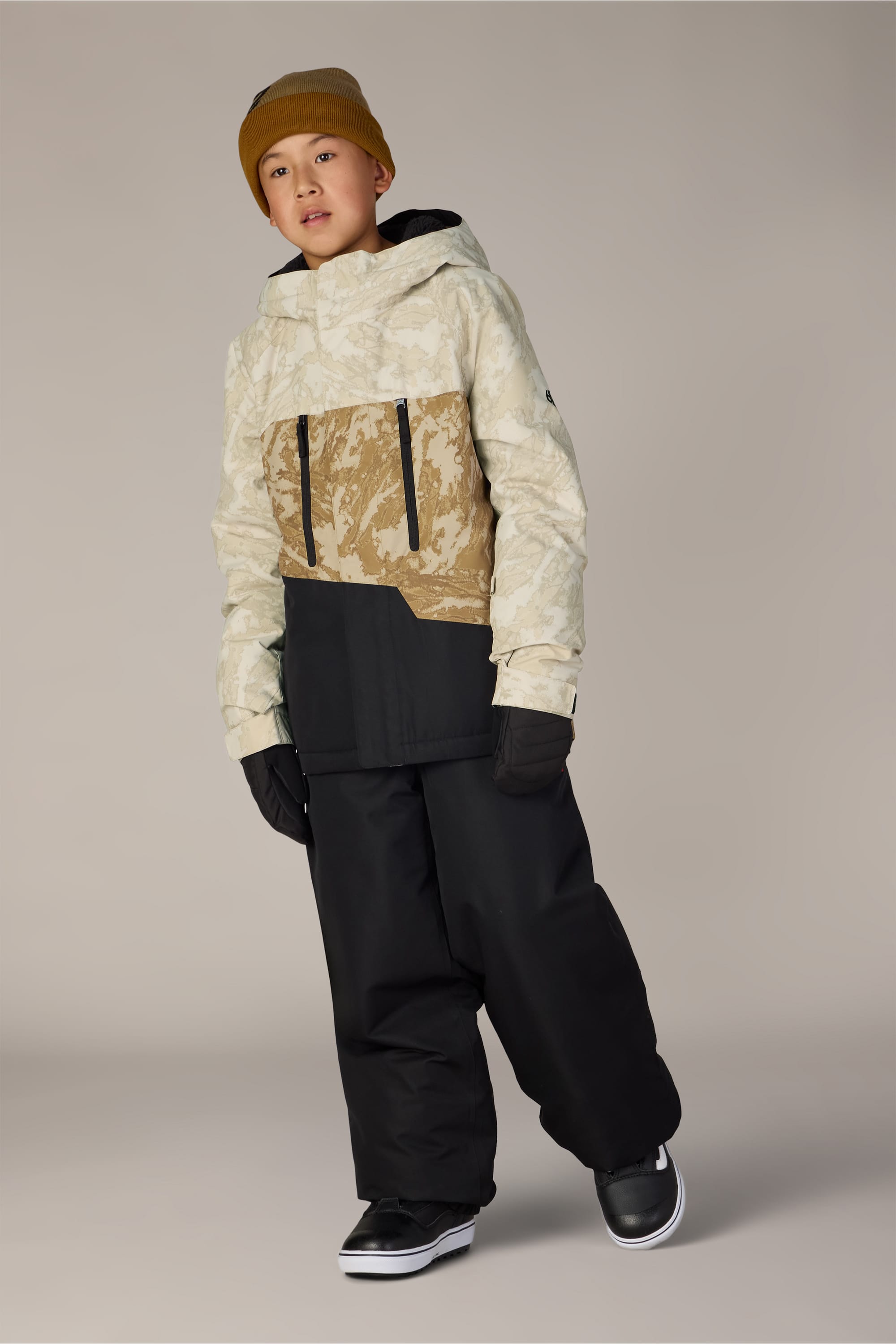 A young person wearing a tan beanie, the 686 Boys’ Geo™ Insulated Jacket in beige and tan camouflage, black snow pants, black gloves, and black shoes stands against a plain background.
