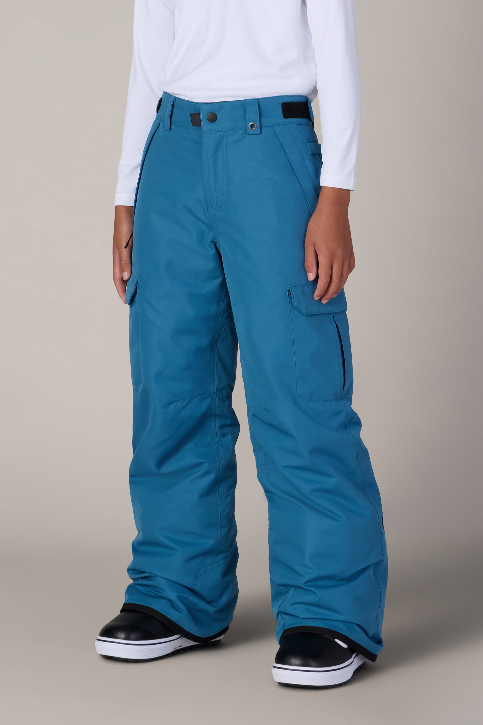 A person wearing 686 Boys Infinity™ Cargo Insulated Pants by 686, a white long-sleeve shirt, and black snow boots stands against a plain light background.
