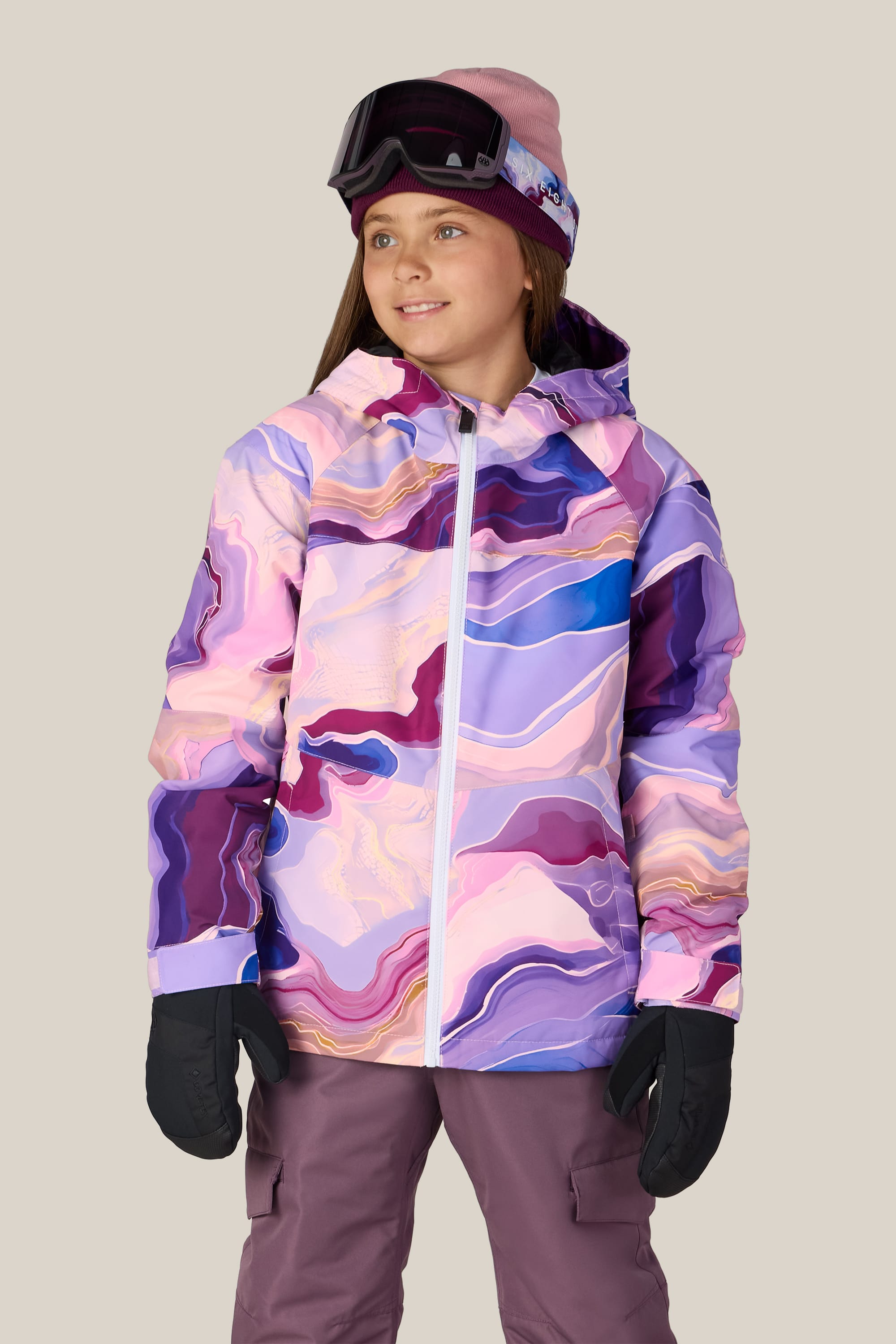 A girl stands against a light background, looking slightly to the side, wearing the 686 Girls’ Athena™ Insulated Jacket by 686 in a pink and purple pattern, paired with purple snow pants, black gloves, a pink beanie, and ski goggles.