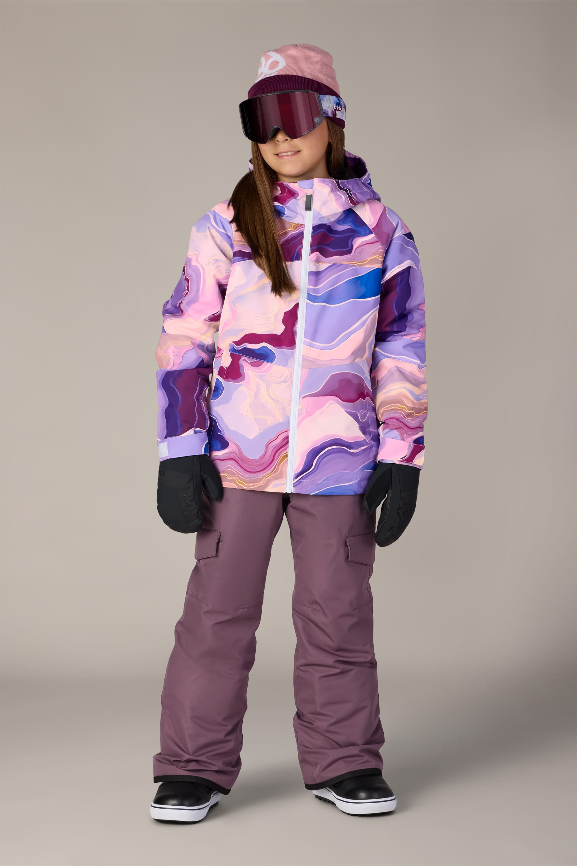 A girl wearing the 686 Girls’ Athena™ Insulated Jacket by 686, purple snow pants, black gloves, ski goggles, and a beanie stands indoors, smiling and looking slightly to the side.