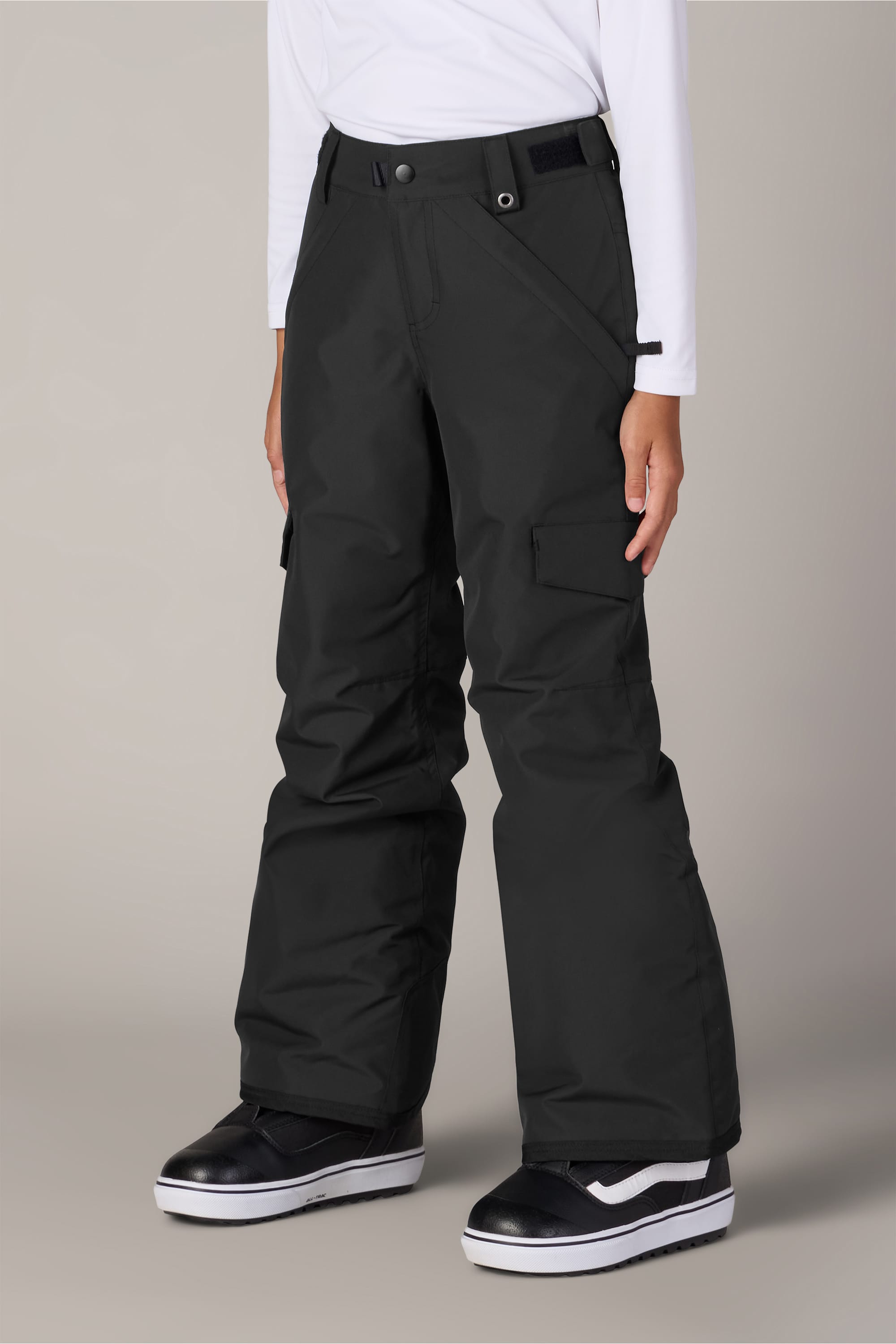 A person is wearing the 686 Girls’ Lola Insulated Pant by 686 in black, paired with a long-sleeve white shirt and black-and-white sneakers, standing against a plain gray background.