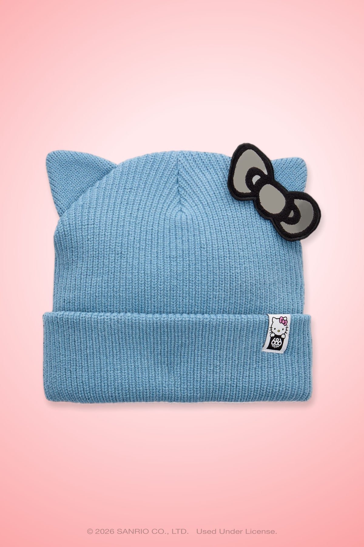 The 686 Women’s Hello Kitty® Knit Beanie by 686 features a light blue knit design with playful cat ears, a gray bow on one side, and a small Hello Kitty tag on the folded brim. The background is a gradient pink.