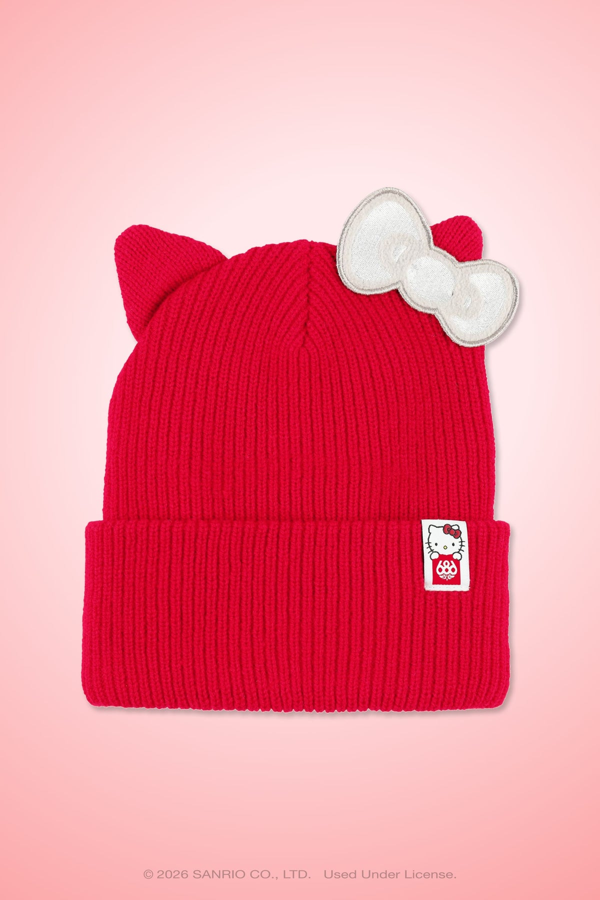 The 686 Women’s Hello Kitty® Knit Beanie by 686 features a red knit design with cat ears, a white bow on one ear, and a small Hello Kitty tag on the brim, displayed against a pink background.