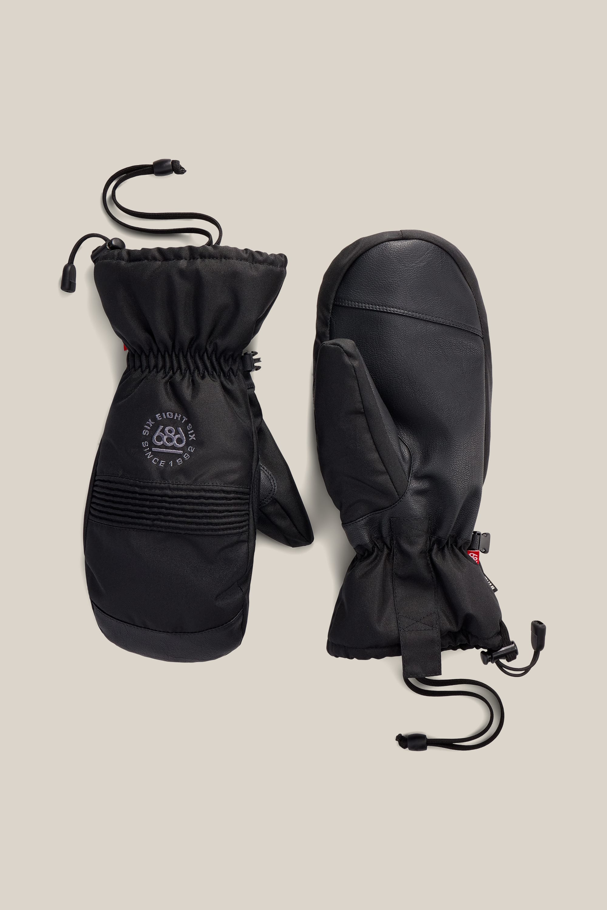 A pair of 686 Lander™ Mitts in black, featuring wrist cinches and elastic cords, is displayed on a neutral background. One mitten shows the circular 686 logo, while the other reveals the palm side.