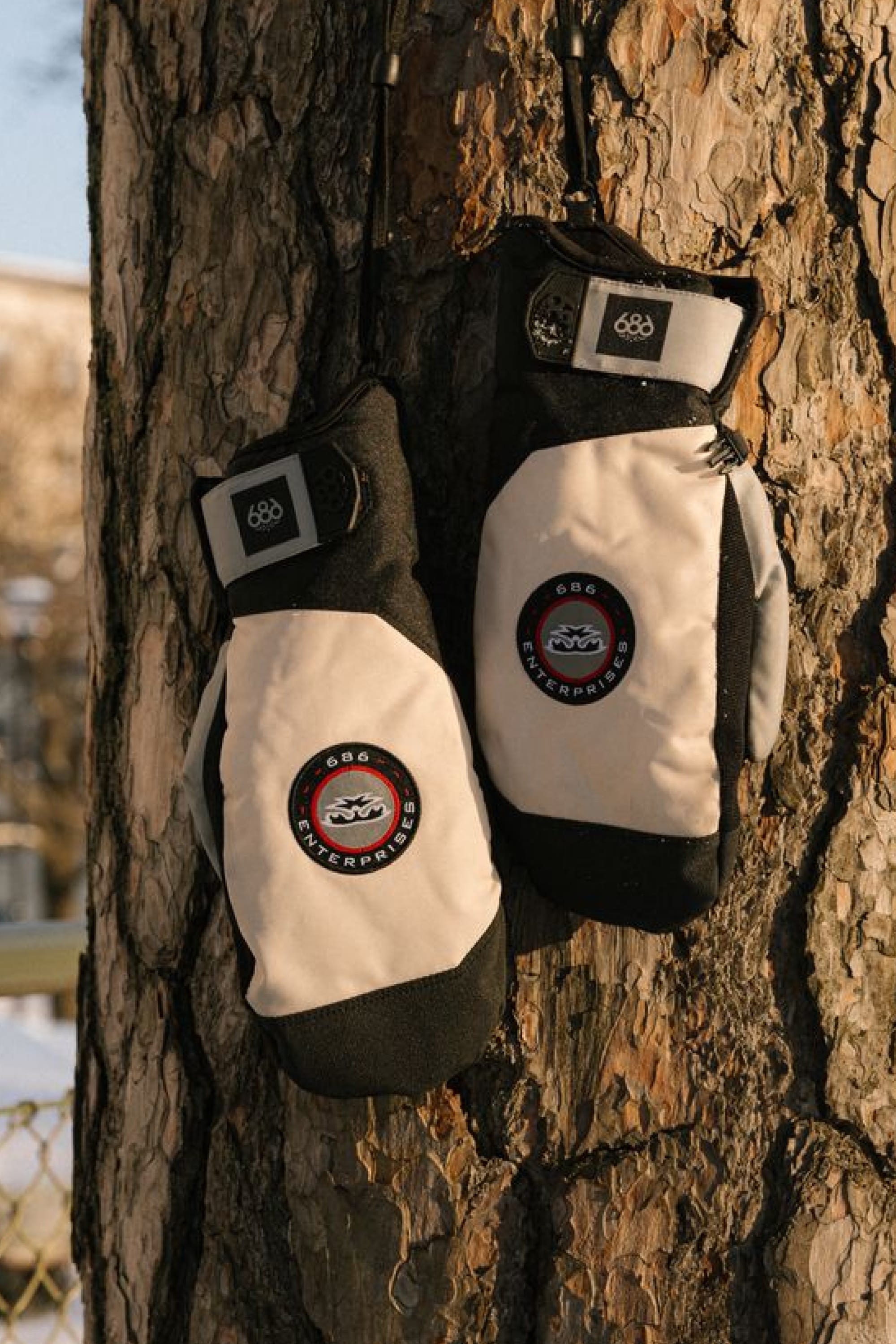 A pair of 686 Men’s Primer Mitts in black and white, featuring circular 686 logos, hang on a tree trunk outdoors as sunlight casts warm shadows across the textured bark.