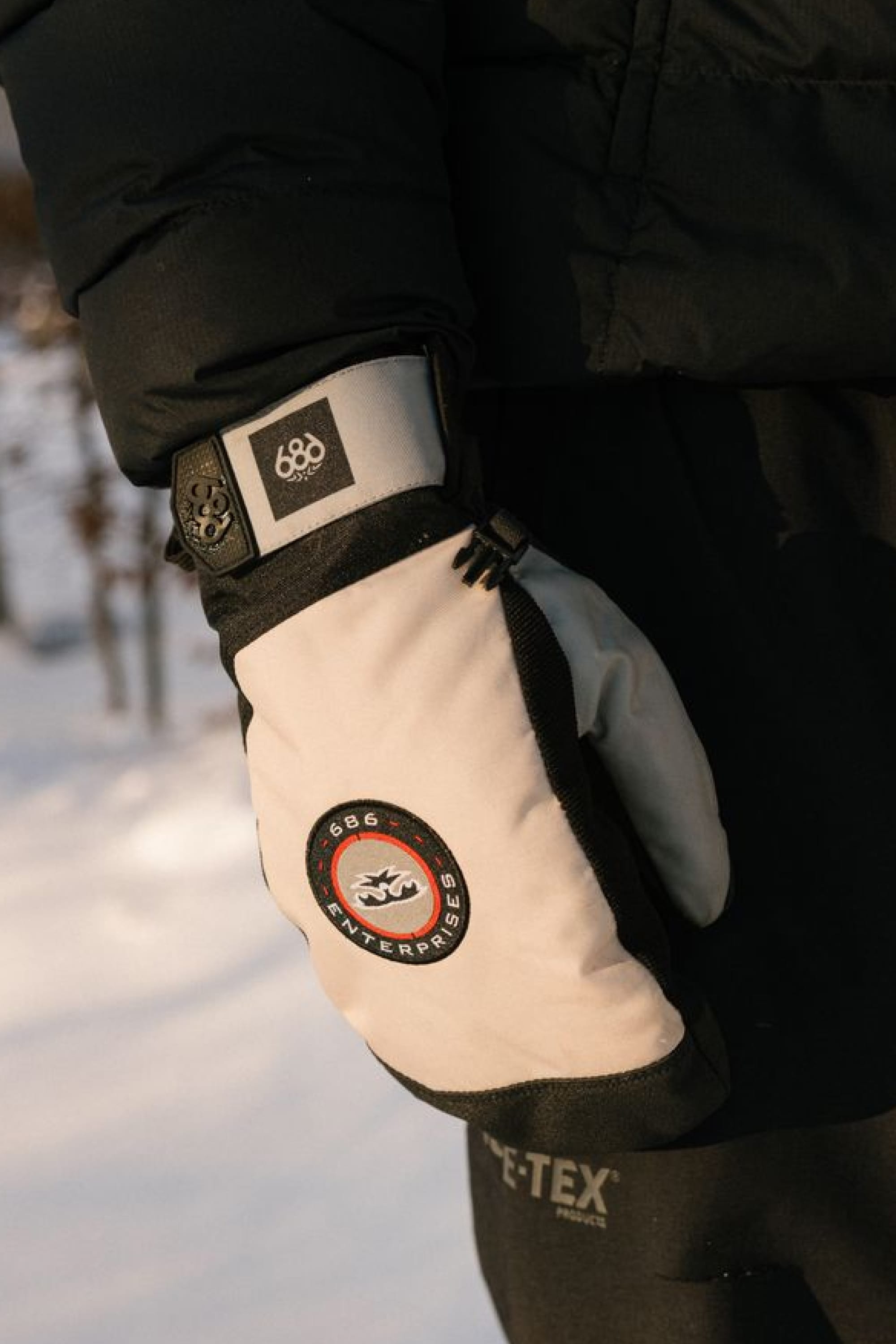 A person stands outdoors in the snow wearing a black jacket and 686 Men’s Primer Mitts by 686.