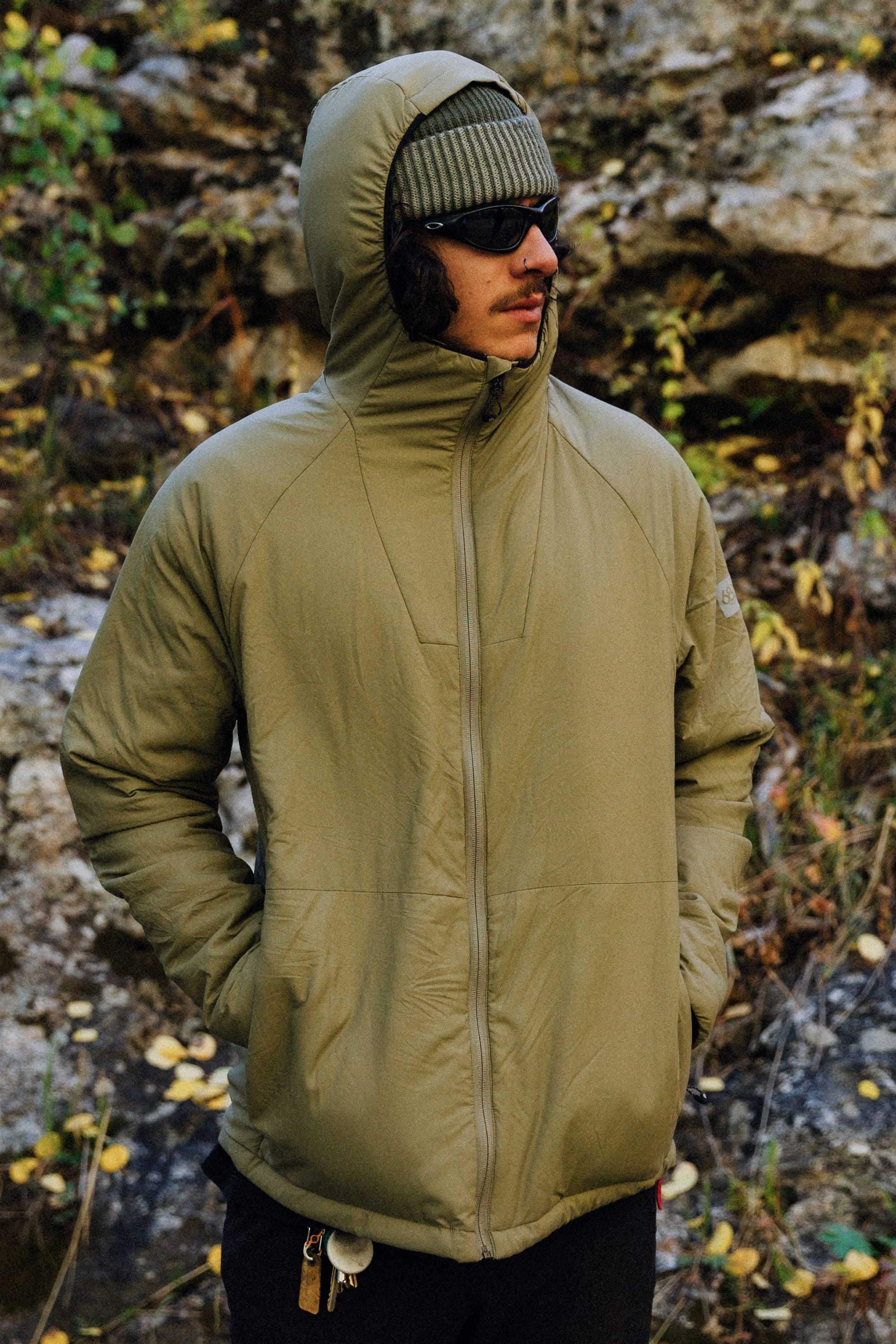 A person wearing the 686 Mens PrimaLoft® Source Jacket in olive, dark sunglasses, and a ribbed beanie stands outdoors with hands in their pockets against a rocky backdrop with some greenery.