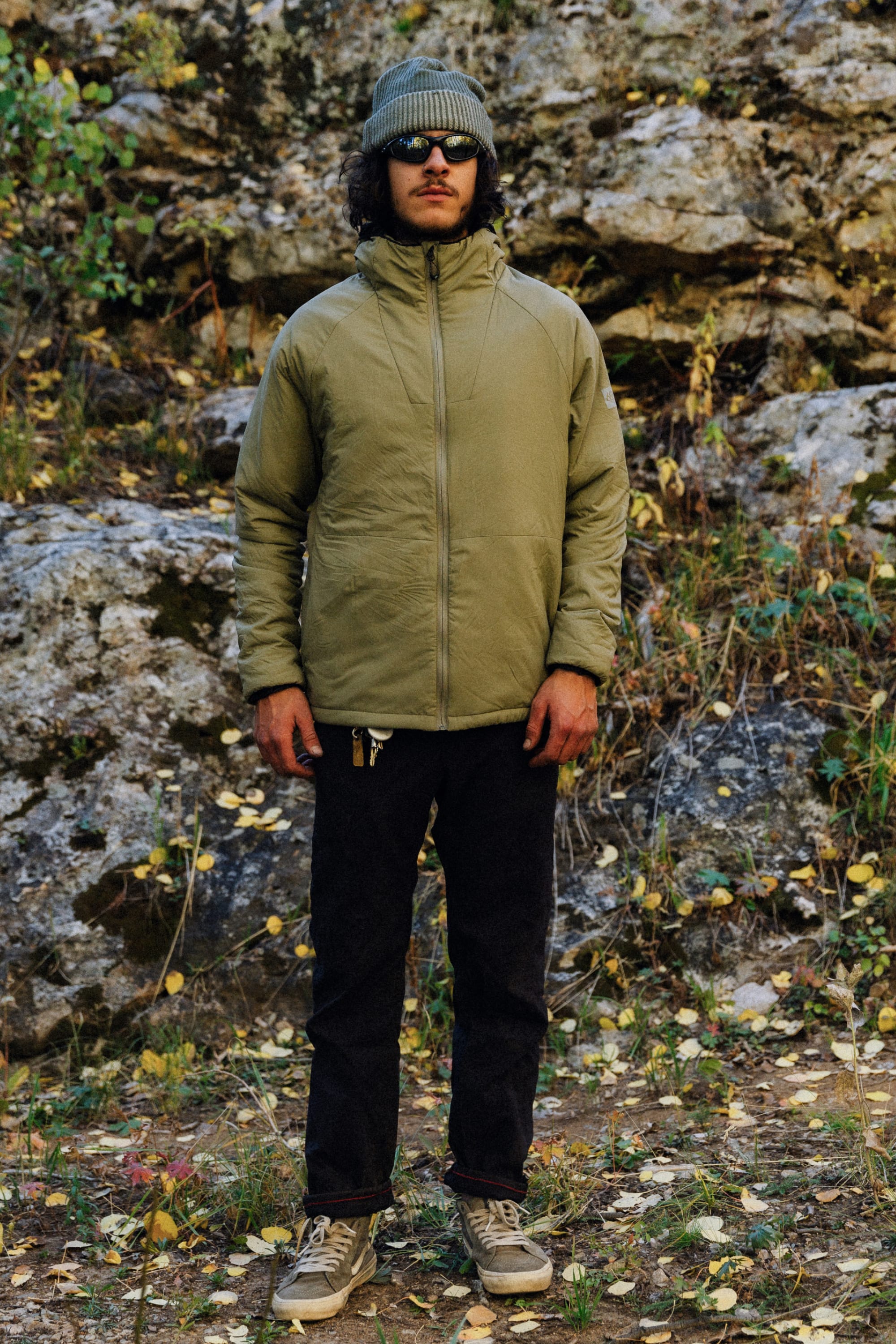 A person in a green beanie, sunglasses, black pants, sneakers, and the 686 Mens PrimaLoft® Source Jacket stands on a rocky path with leaves, greenery, and large rocks in the background.