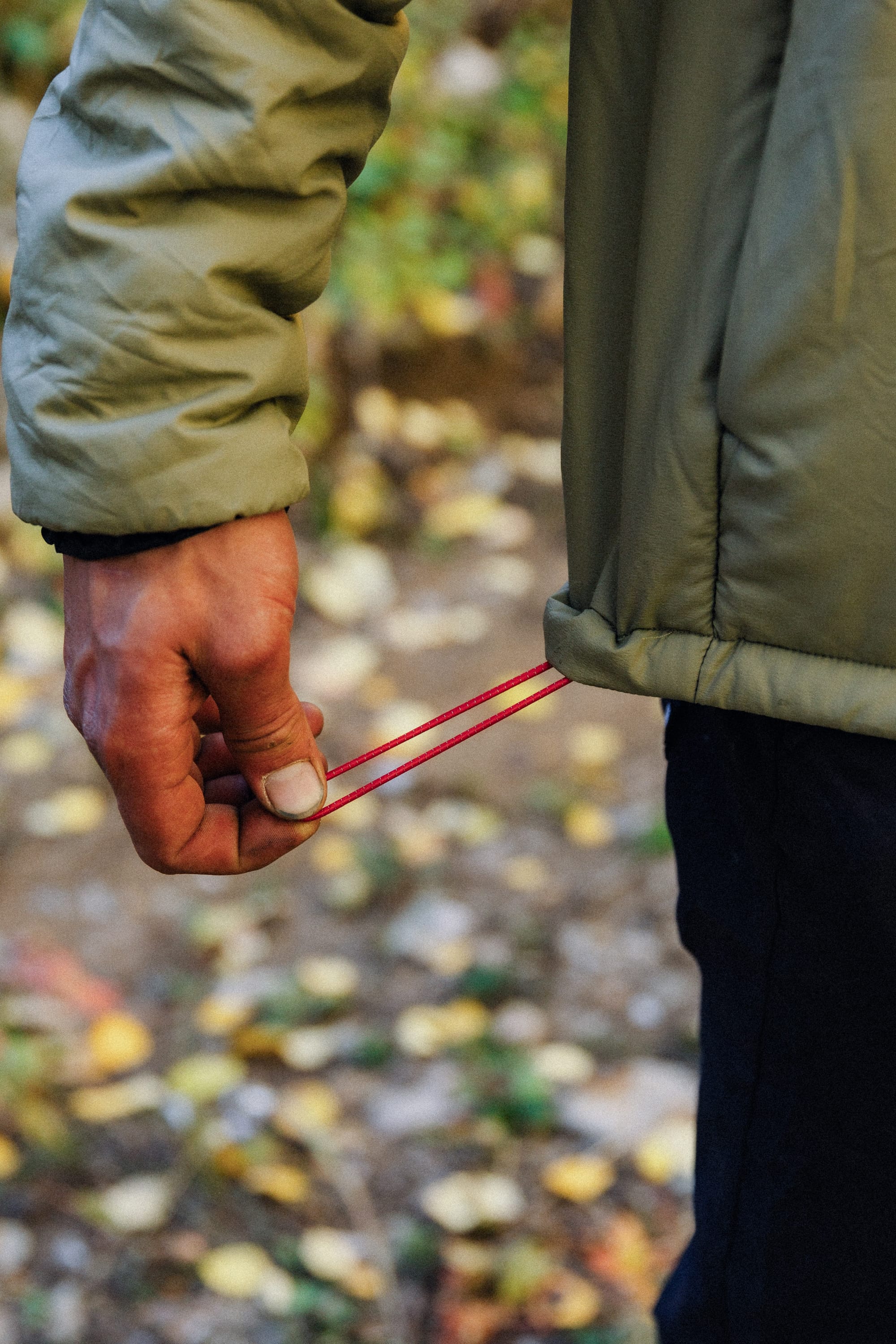 A person in a 686 Mens PrimaLoft® Source Jacket adjusts the red hem drawstring. Fallen leaves cover the outdoor setting.