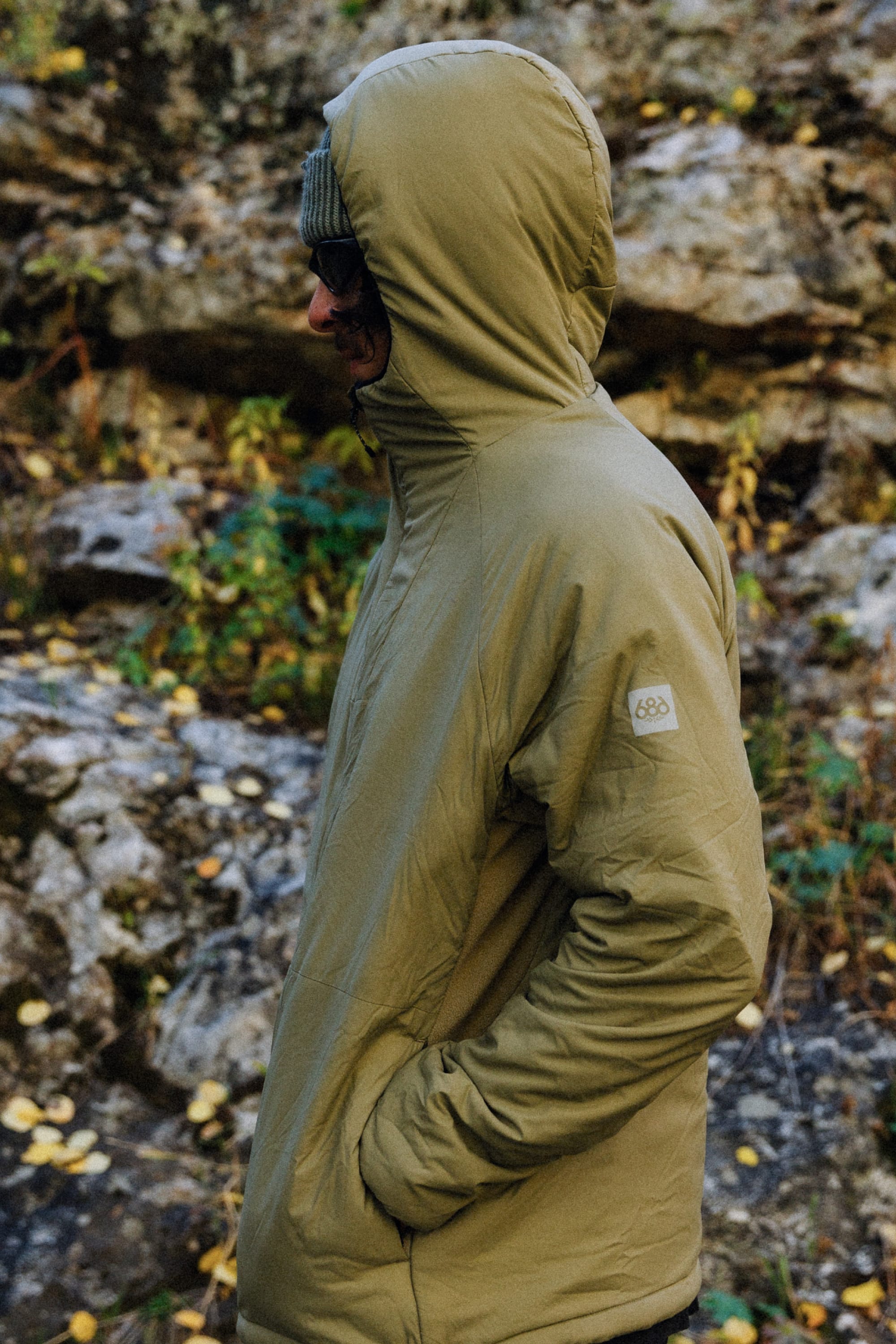 A person in the 686 Mens PrimaLoft® Source Jacket in olive green and dark sunglasses stands sideways outdoors near rocky terrain with scattered yellow leaves.