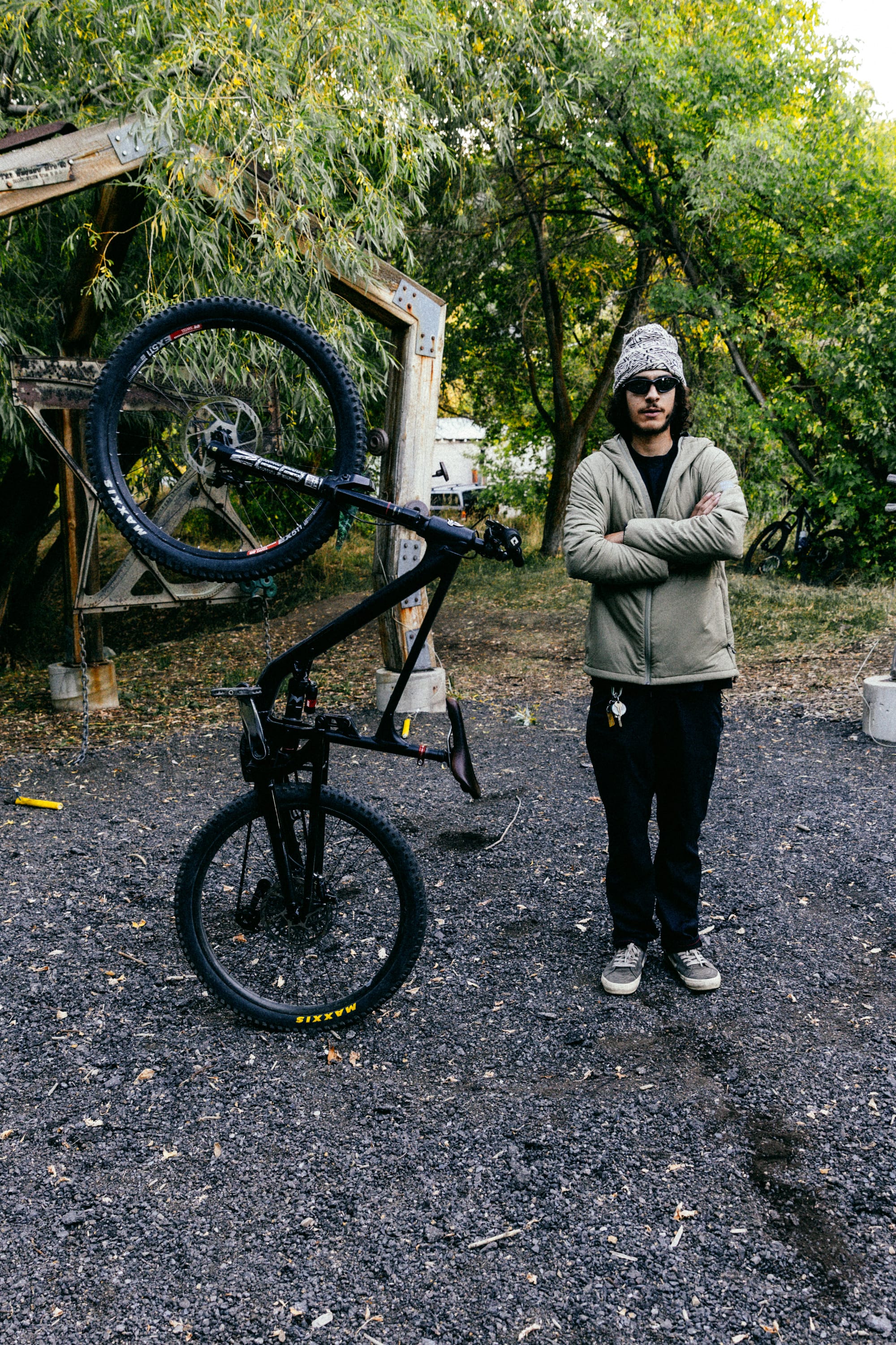 Wearing the 686 Mens PrimaLoft® Source Jacket, a person in sunglasses and a beanie stands with arms crossed by a black bicycle balanced on its rear wheel outdoors among trees and gravel.