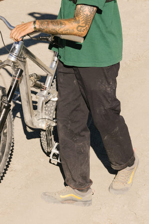 A person with tattooed arms, wearing a green shirt, 686 Men's Everywhere® Pant - Straight Fit from 686, and gray shoes with yellow accents walks a mountain bike on a dusty path.