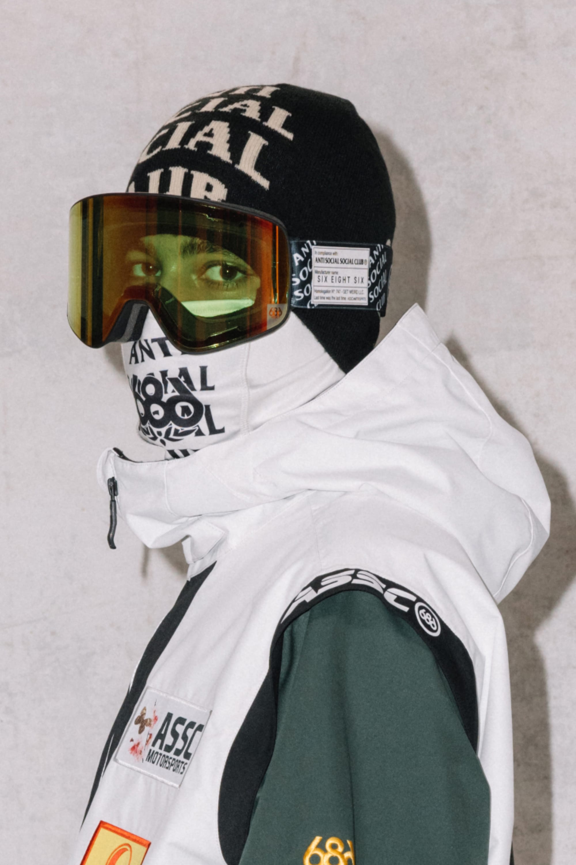 A person wearing a black beanie, 686 SMARTY® Magnetic Goggle by 686 with interchangeable lenses, a white face covering with text, and a white and green patch-adorned winter jacket stands against a plain light background.