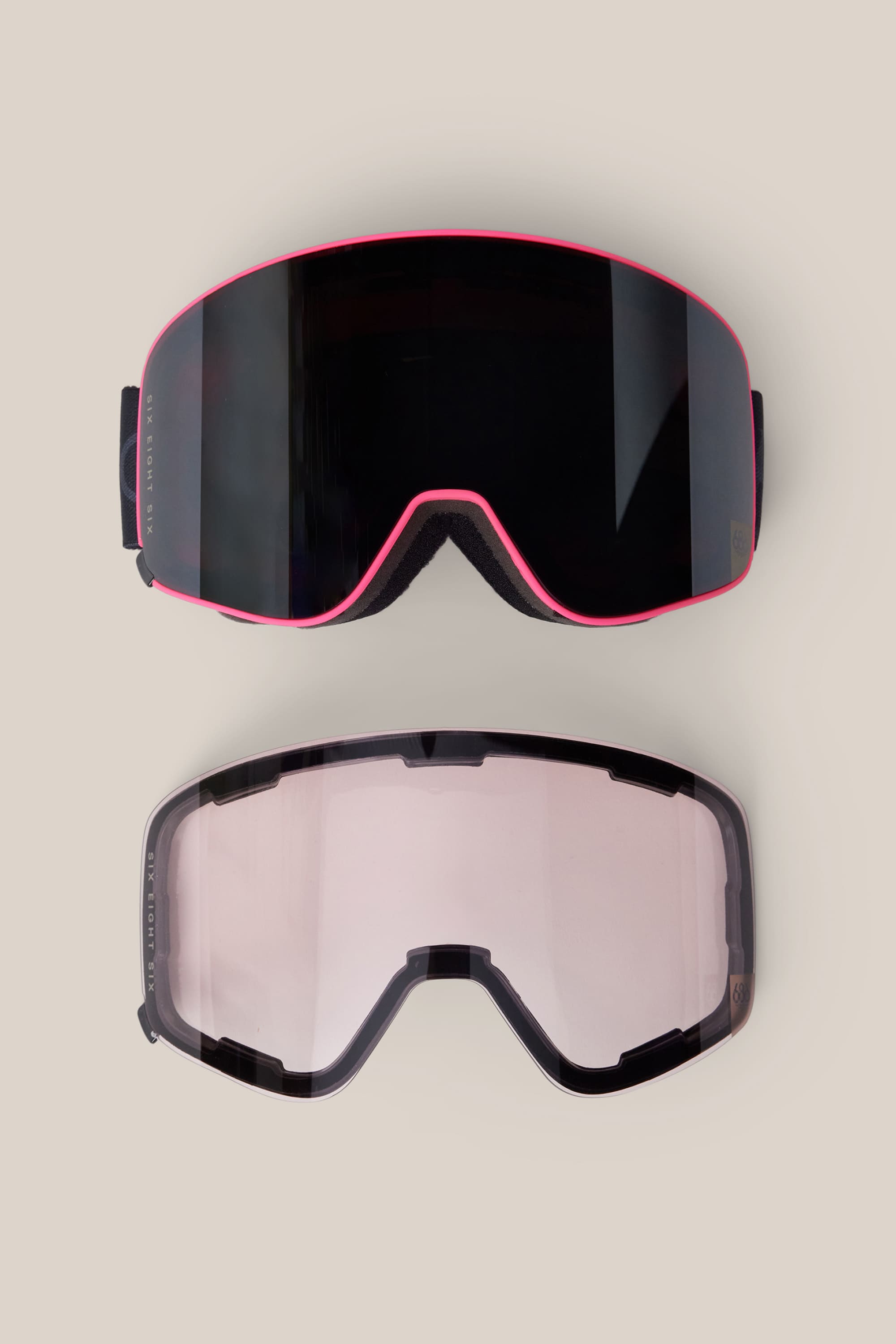 Two interchangeable lenses for the 686 SMARTY® Magnetic Goggle by 686 are displayed on a beige background: one is dark and reflective with a pink rim, while the other is clear with a light tint and features a black rim.