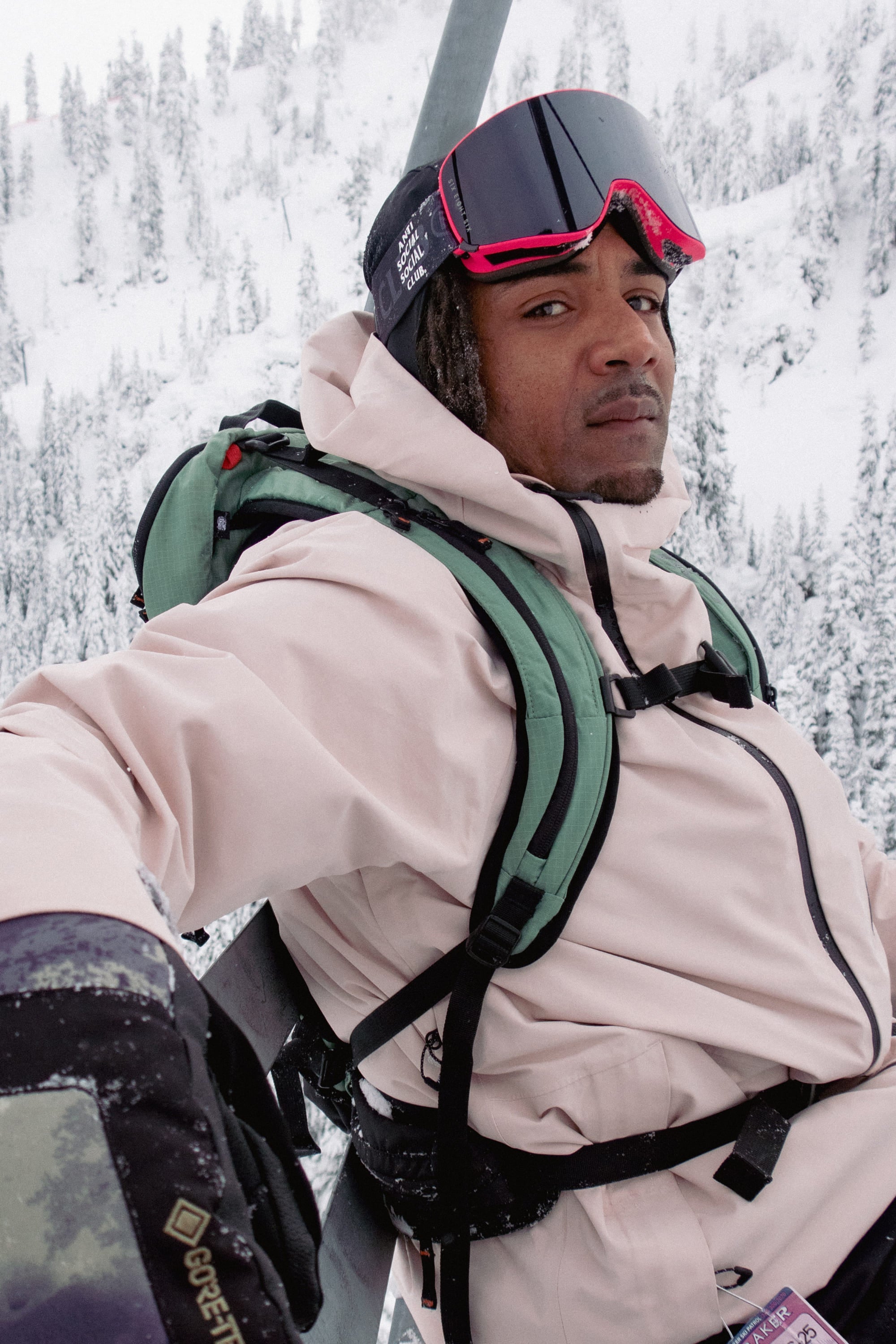 Wearing a light pink jacket and 686 SMARTY® Magnetic Goggles by 686, a person with a green backpack and gloves sits on a ski lift amid snowy mountains, with snow-covered trees in the background.