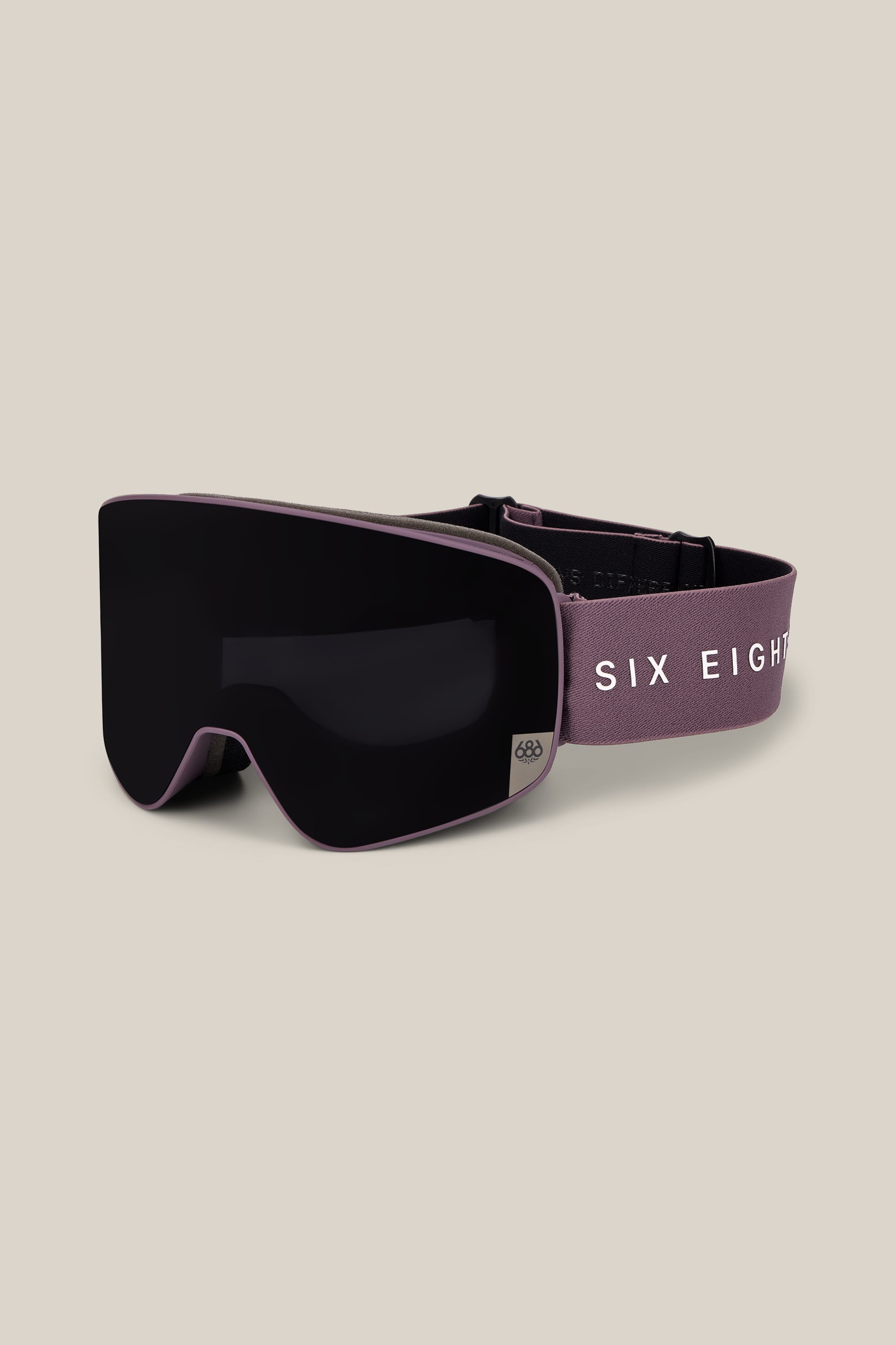 The 686 SMARTY® Magnetic Goggle features a matte black frame, a wide dark lens, and a mauve strap with “SIX EIGHT” in white lettering, showcased against a beige background.