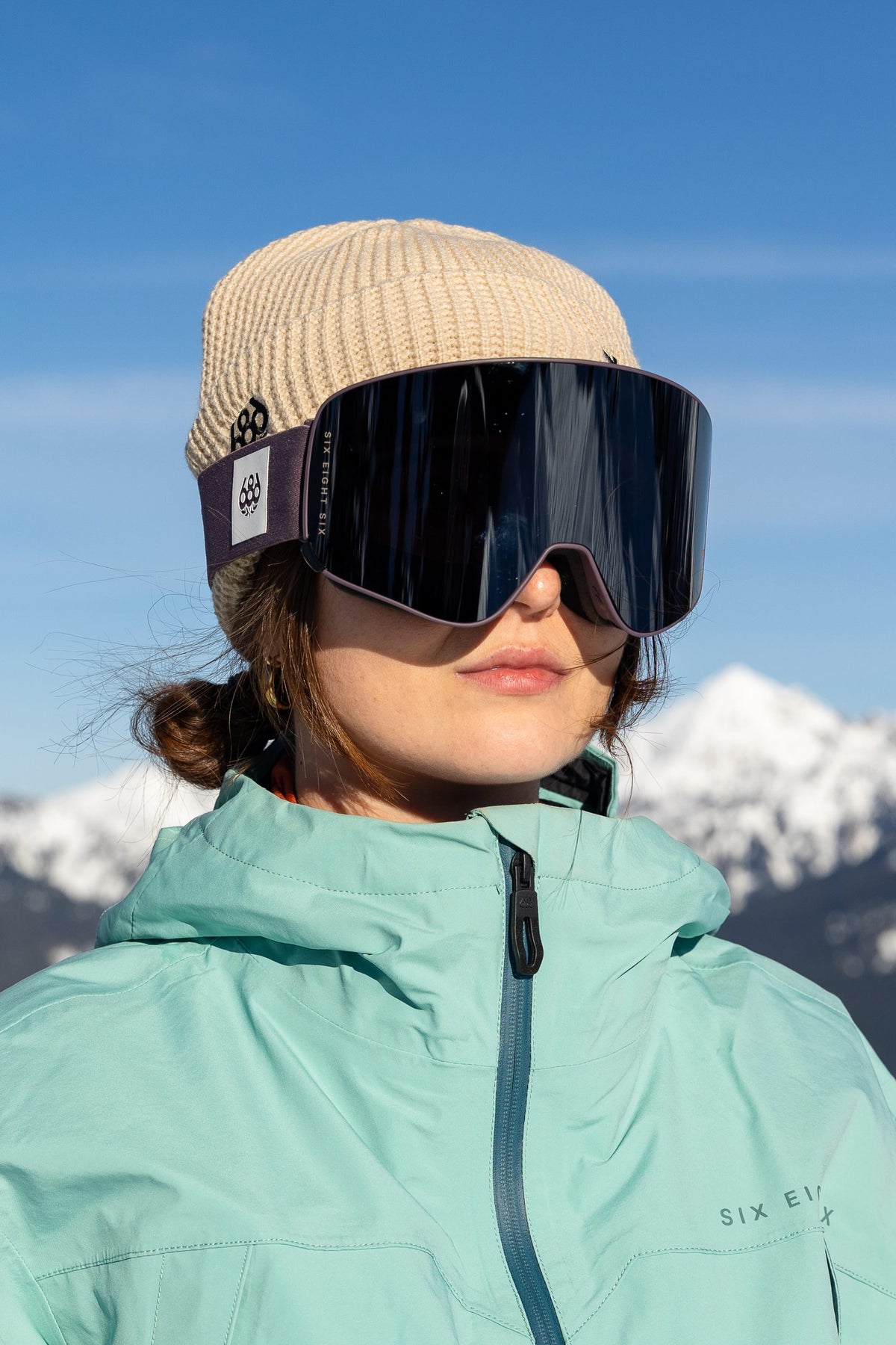 A person wearing a beige knit hat, 686 SMARTY® Magnetic Goggles by 686, and a light blue jacket stands outdoors with snowy mountains and a blue sky in the background.
