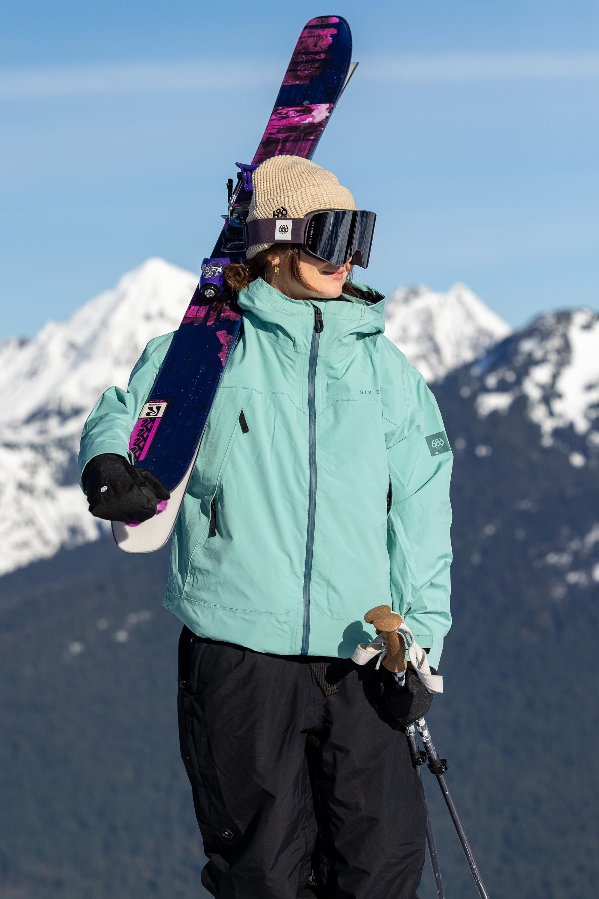 Wearing the 686 SMARTY® Magnetic Goggle by 686, a person in a light blue jacket and tan beanie holds skis on their shoulder while standing on a snowy mountain with peaks visible in the background.