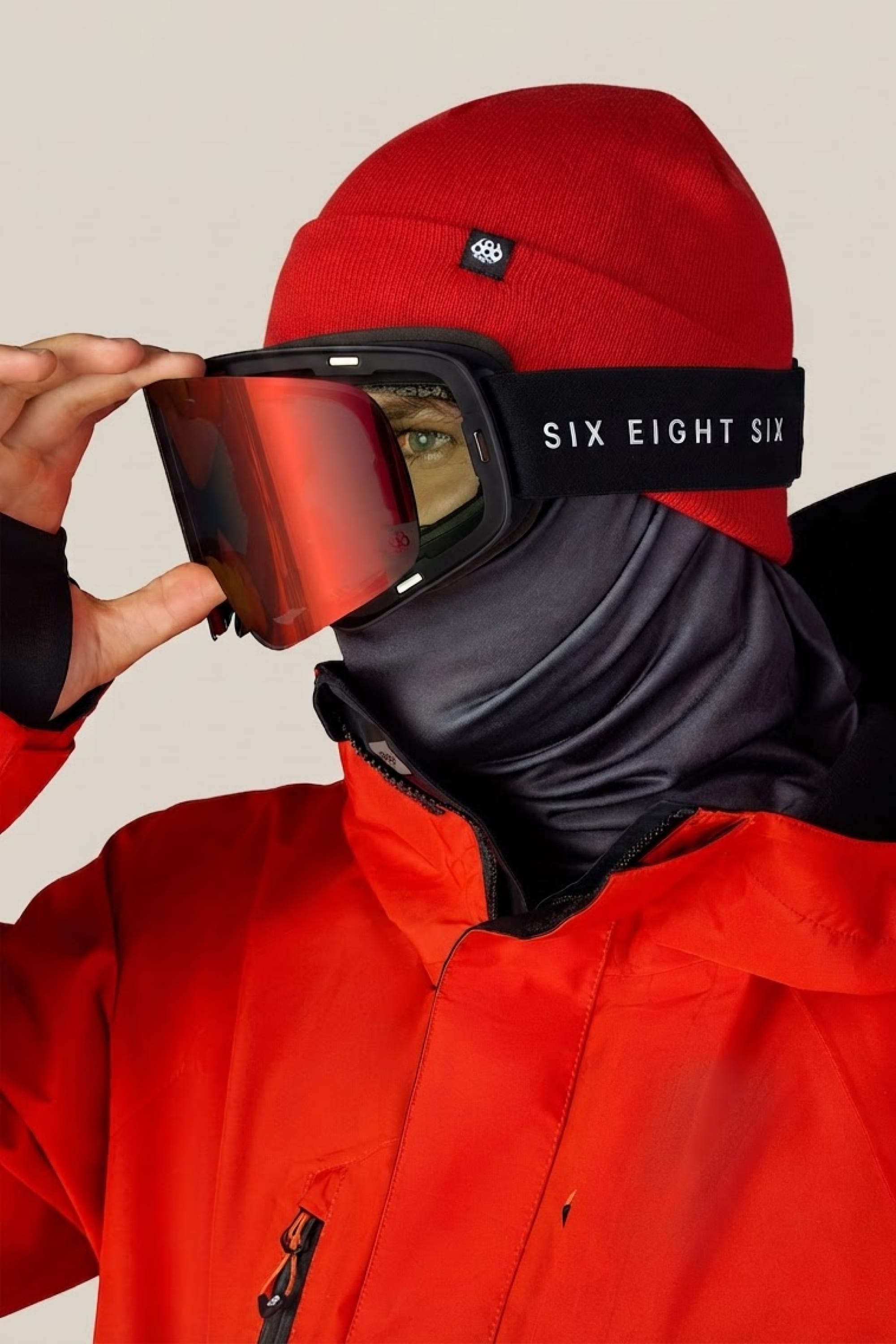 A person in a red jacket and beanie adjusts their 686 SMARTY® Magnetic Goggle, featuring a black face covering and visible 686 branding on the goggle strap.