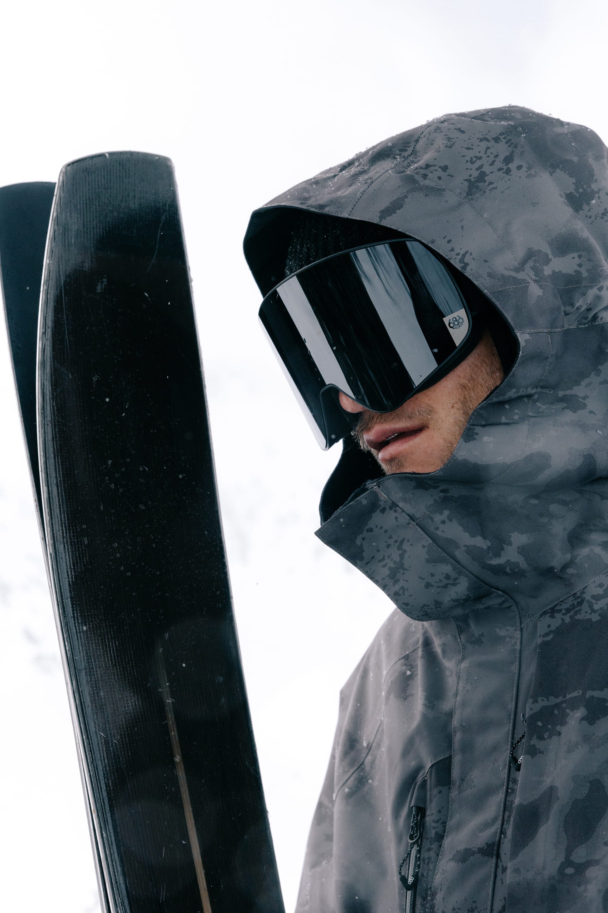 A person in a gray camo-patterned ski jacket and large 686 SMARTY® Magnetic Goggles by 686, holding skis upright against a snowy, overcast background.