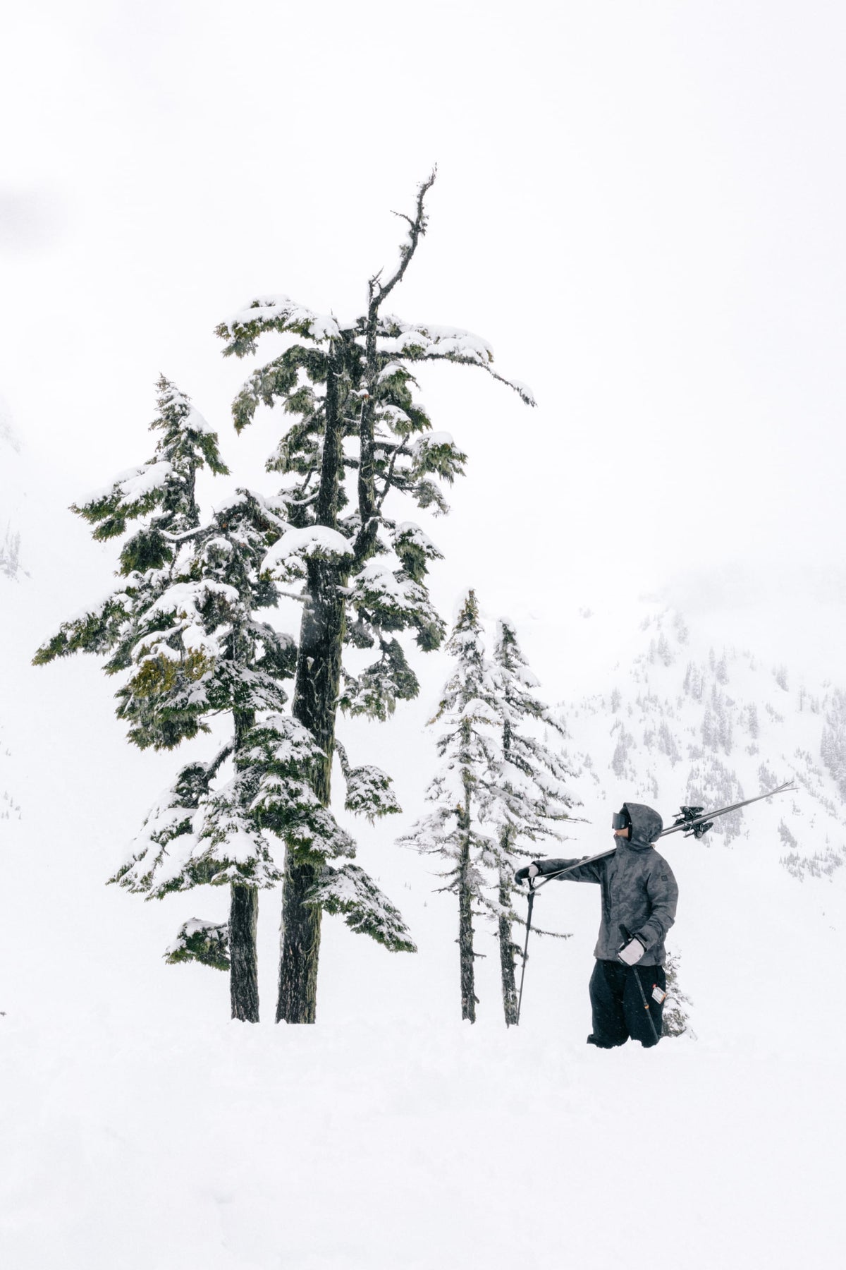 Wearing the 686 SMARTY® Magnetic Goggle by 686, a person in winter clothing stands in deep snow beside snow-covered pine trees, with a snowy mountain landscape in the background.