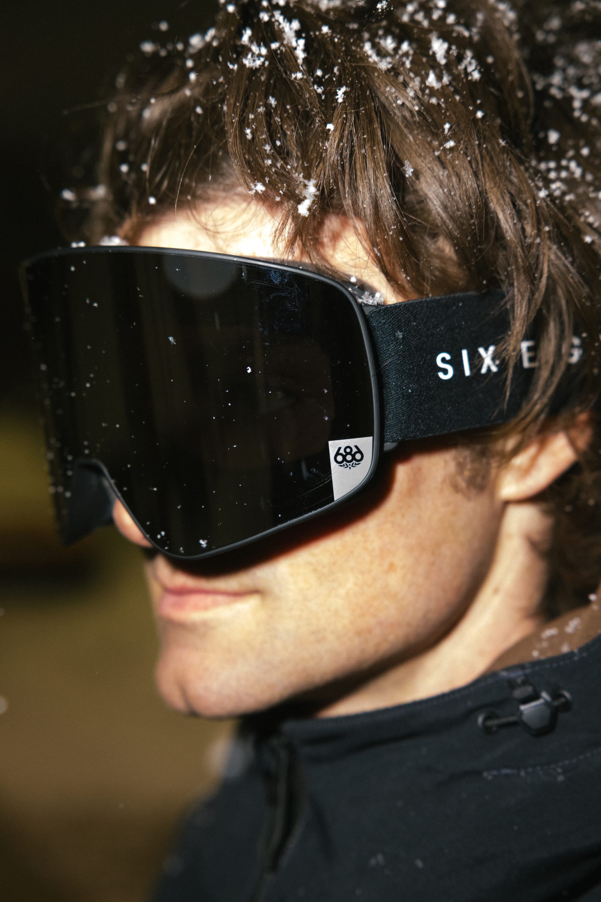 A person wearing the 686 SMARTY® Magnetic Goggle, with the 686 logo visible on the side, dressed in a dark jacket, snowflakes resting on their hair and goggles.