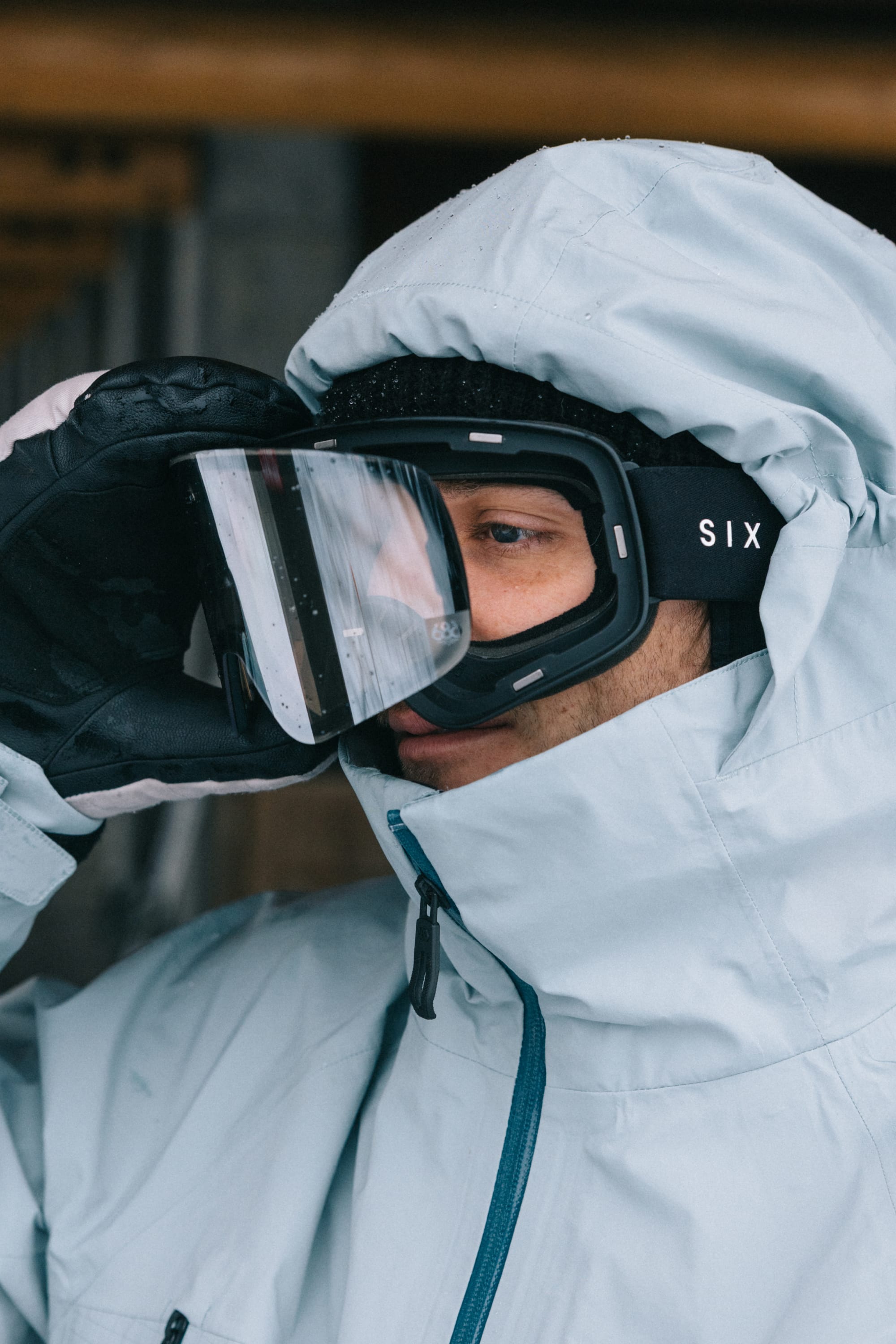 Wearing a light blue winter jacket and black gloves, a person adjusts their 686 SMARTY® Magnetic Goggle, with the word 686 visible on the side strap. Snow or frost is reflected on the goggles.