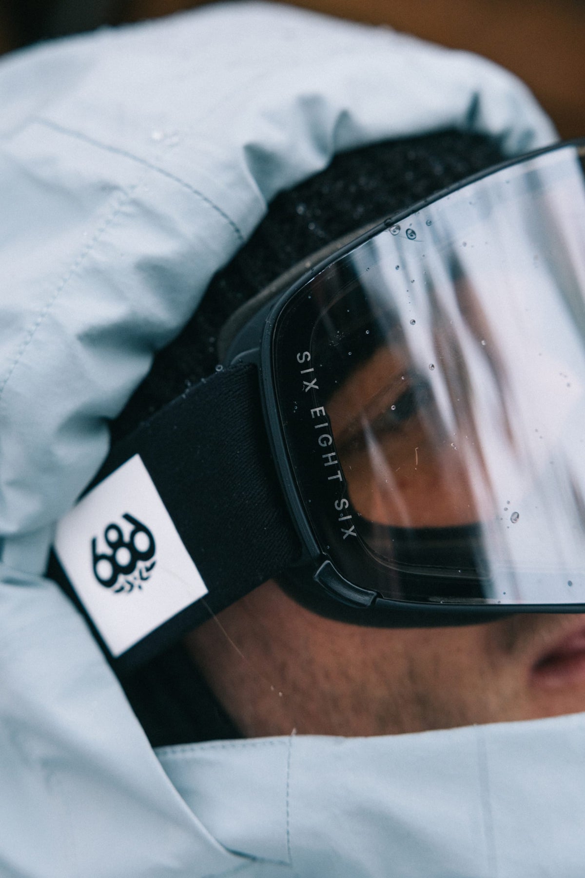 A close-up of a person wearing a light blue hooded jacket, black beanie, and the 686 SMARTY® Magnetic Goggle with “686” branding visible on the strap. The focus is on the goggles and their branded strap.