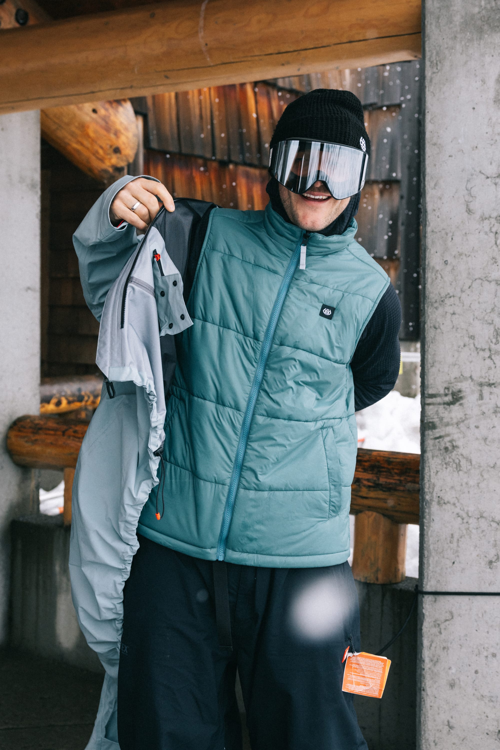 A person wearing a blue puffer vest, black beanie, and the 686 SMARTY® Magnetic Goggle by 686 stands under a wooden structure, holding a light-colored jacket and smiling with snow visible in the background.