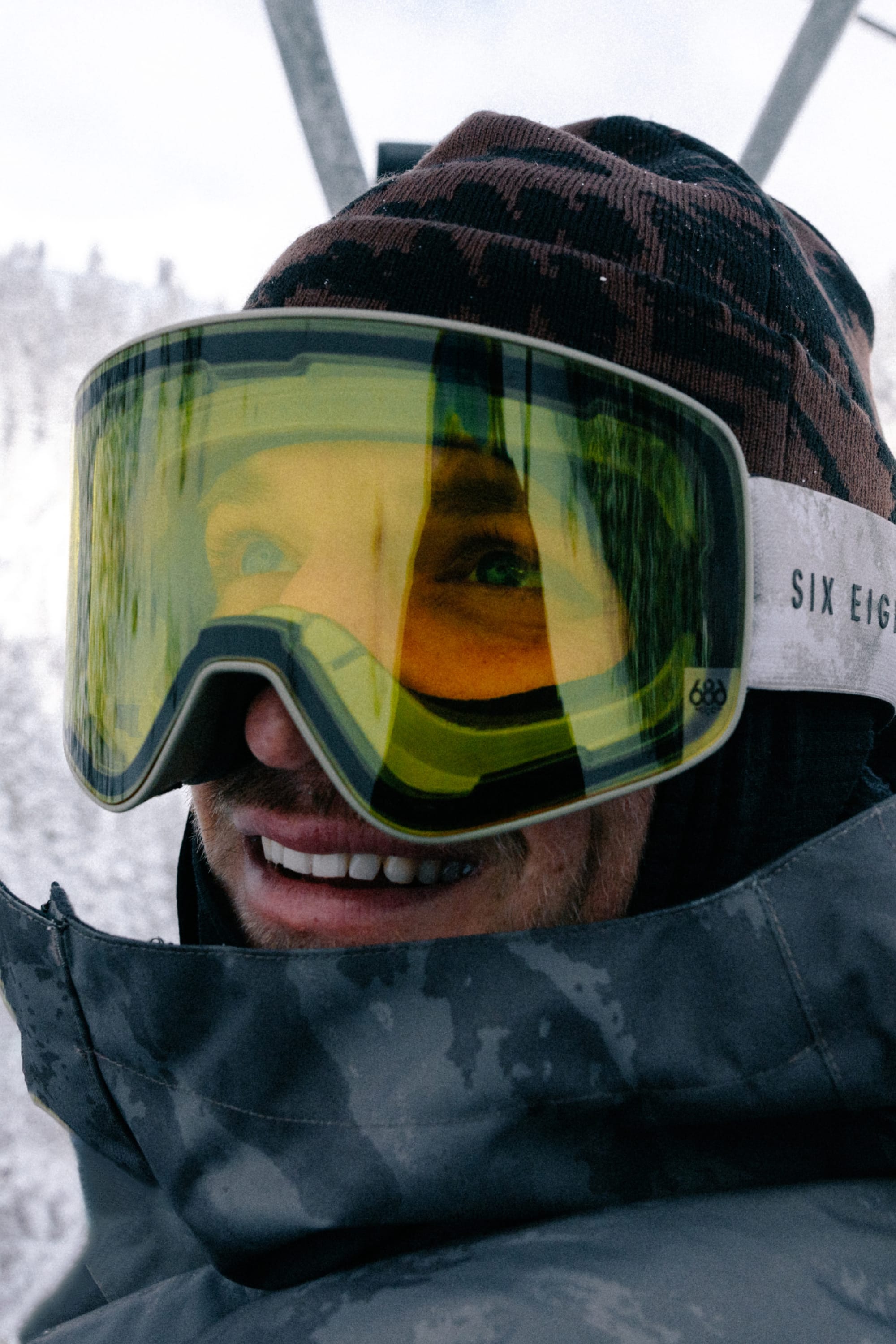 Wearing the 686 SMARTY® Magnetic Goggle by 686, a person in a brown hat and gray jacket smiles on a snowy mountain, with trees and a lift in the background.