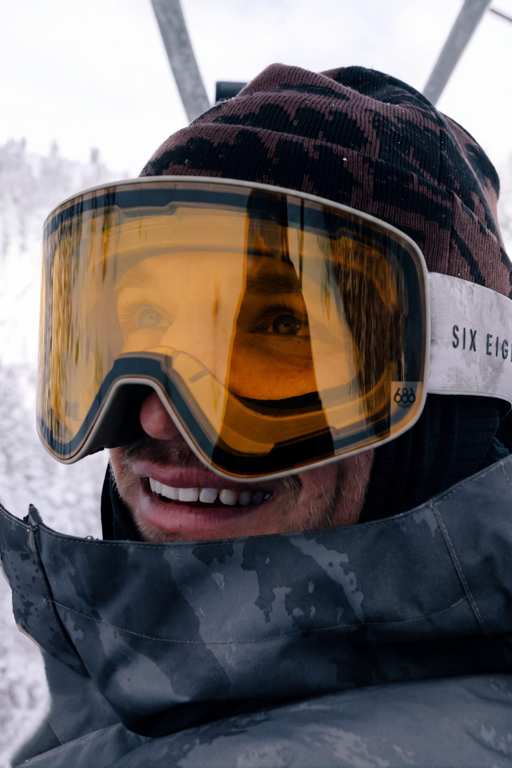 Wearing winter gear and a dark jacket, a person smiles outdoors in front of snowy, blurred trees, their eyes protected by the 686 SMARTY® Magnetic Goggle from 686.