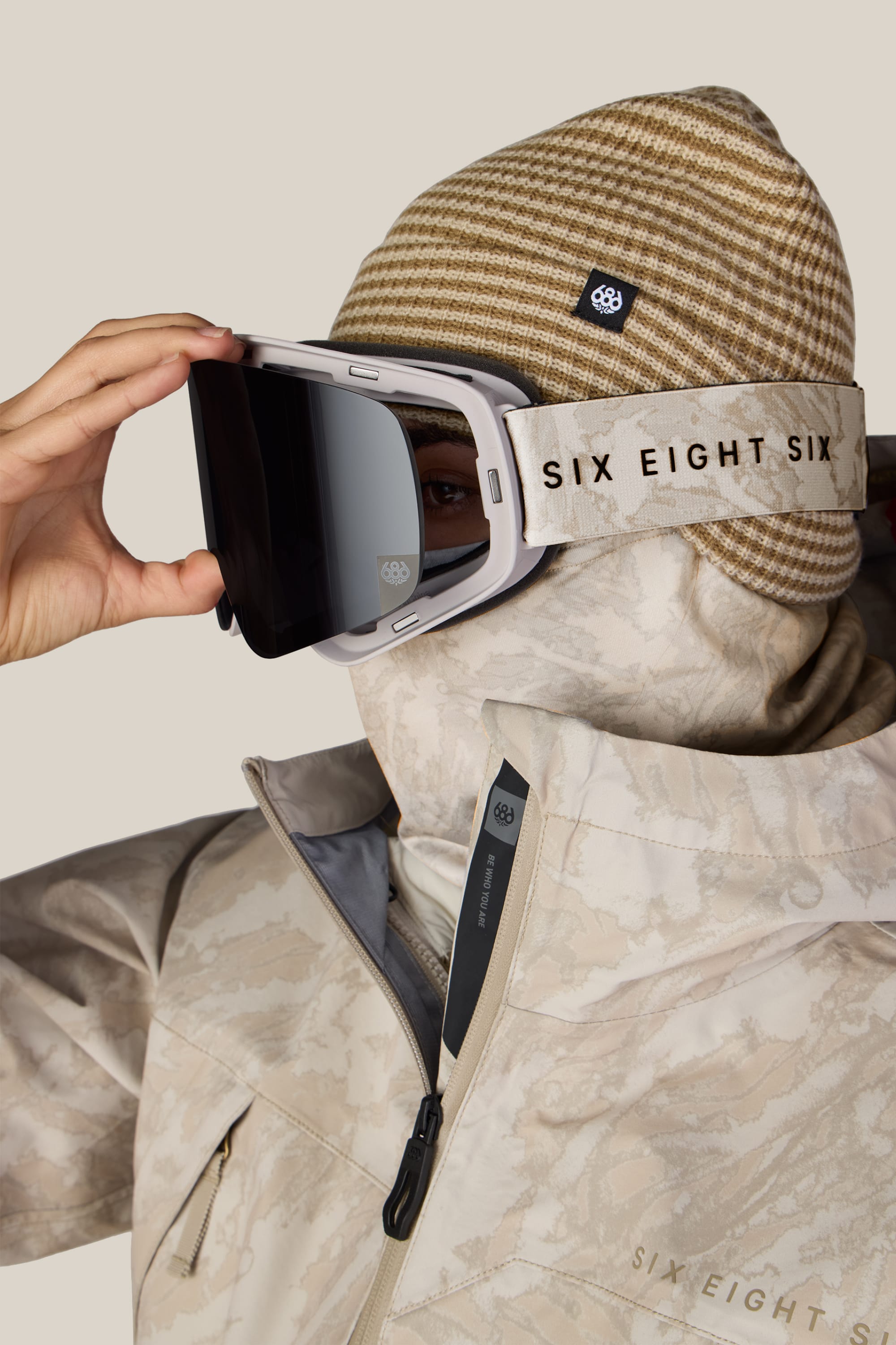 A person is wearing beige patterned winter gear, a brown-striped beanie, and the 686 SMARTY® Magnetic Goggle by 686. They are adjusting their goggles, and their jacket zipper is partially open.
