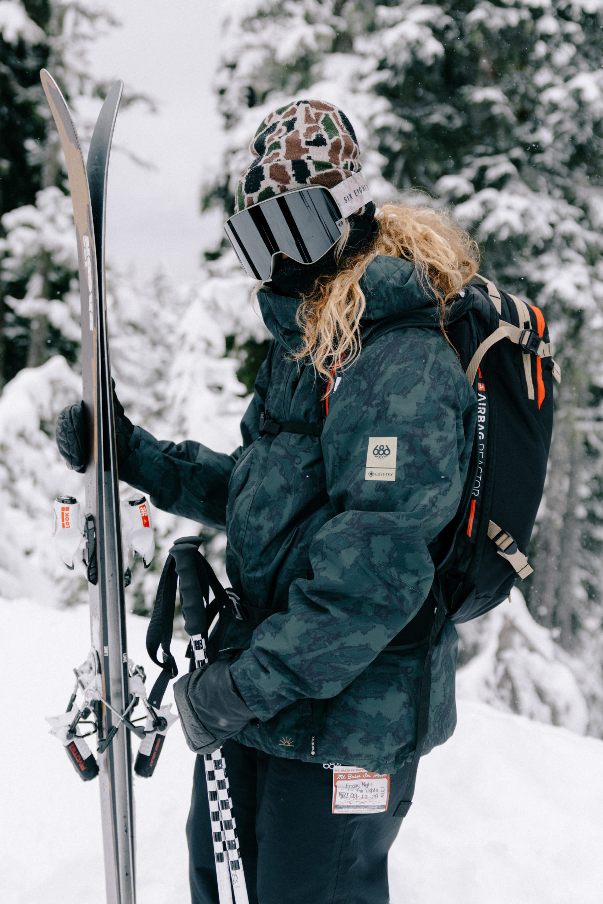 A person in winter gear and a patterned beanie stands in snowy woods, holding skis and poles, with curly hair peeking out beneath a 686 SMARTY® Magnetic Goggle by 686 and a backpack on their back.