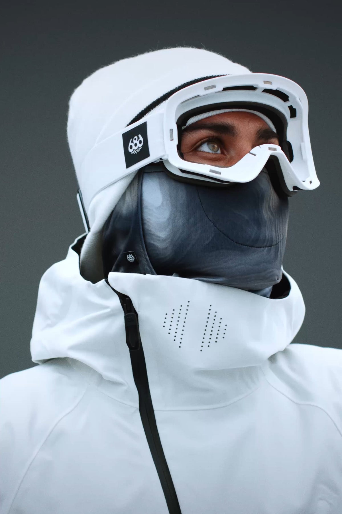 A person wearing a white jacket, a gray face mask, and 686 SMARTY® Magnetic Goggles by 686 looks upward. The plain, dark background emphasizes the winter or snow sports attire.