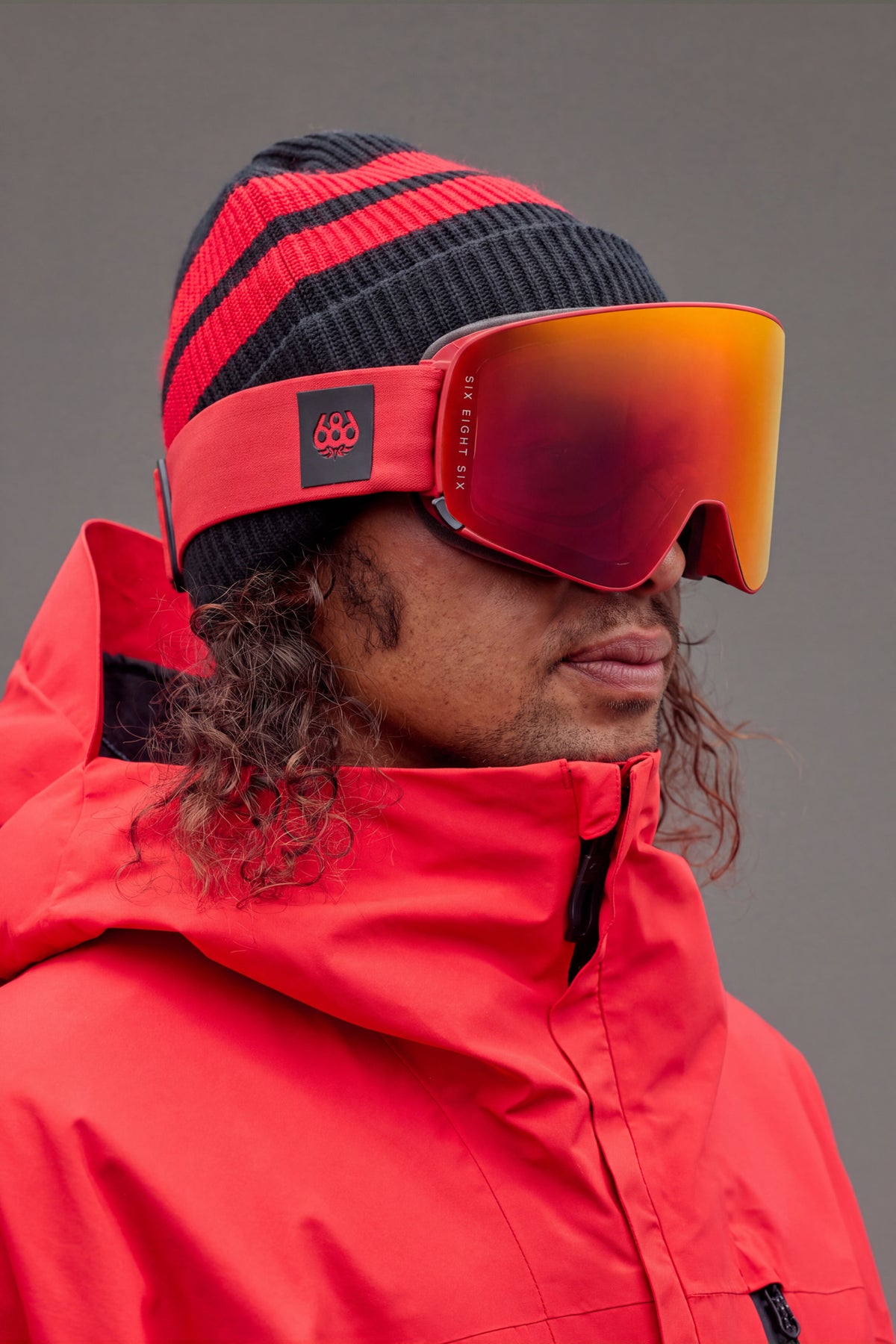 A person in a red jacket and striped beanie wears the 686 SMARTY® Magnetic Goggle by 686, looking to the side against a plain background.