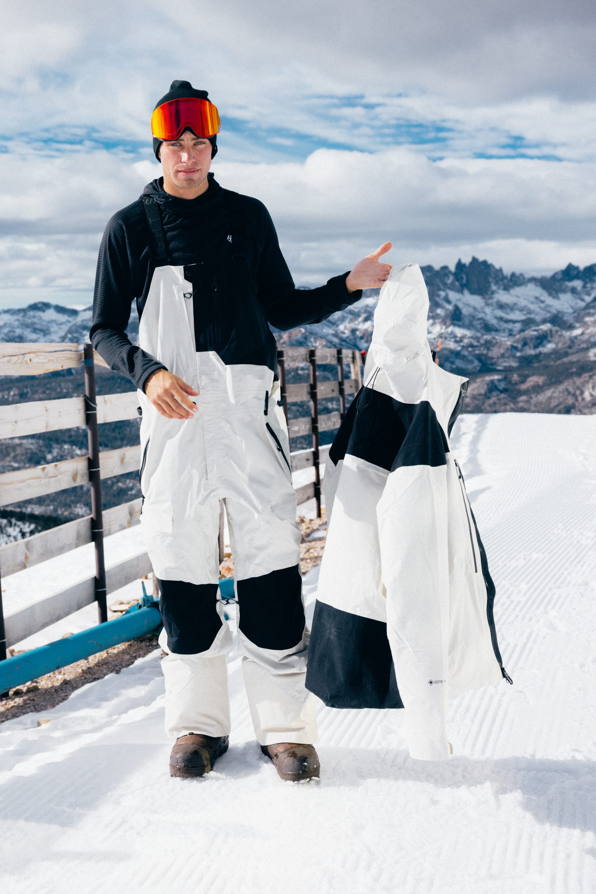 Wearing the 686 SMARTY® Magnetic Goggle by 686, a person dressed in white and black ski gear stands on a snowy mountain, holding a matching jacket. They also wear a beanie, with mountains and a wooden fence in the background.