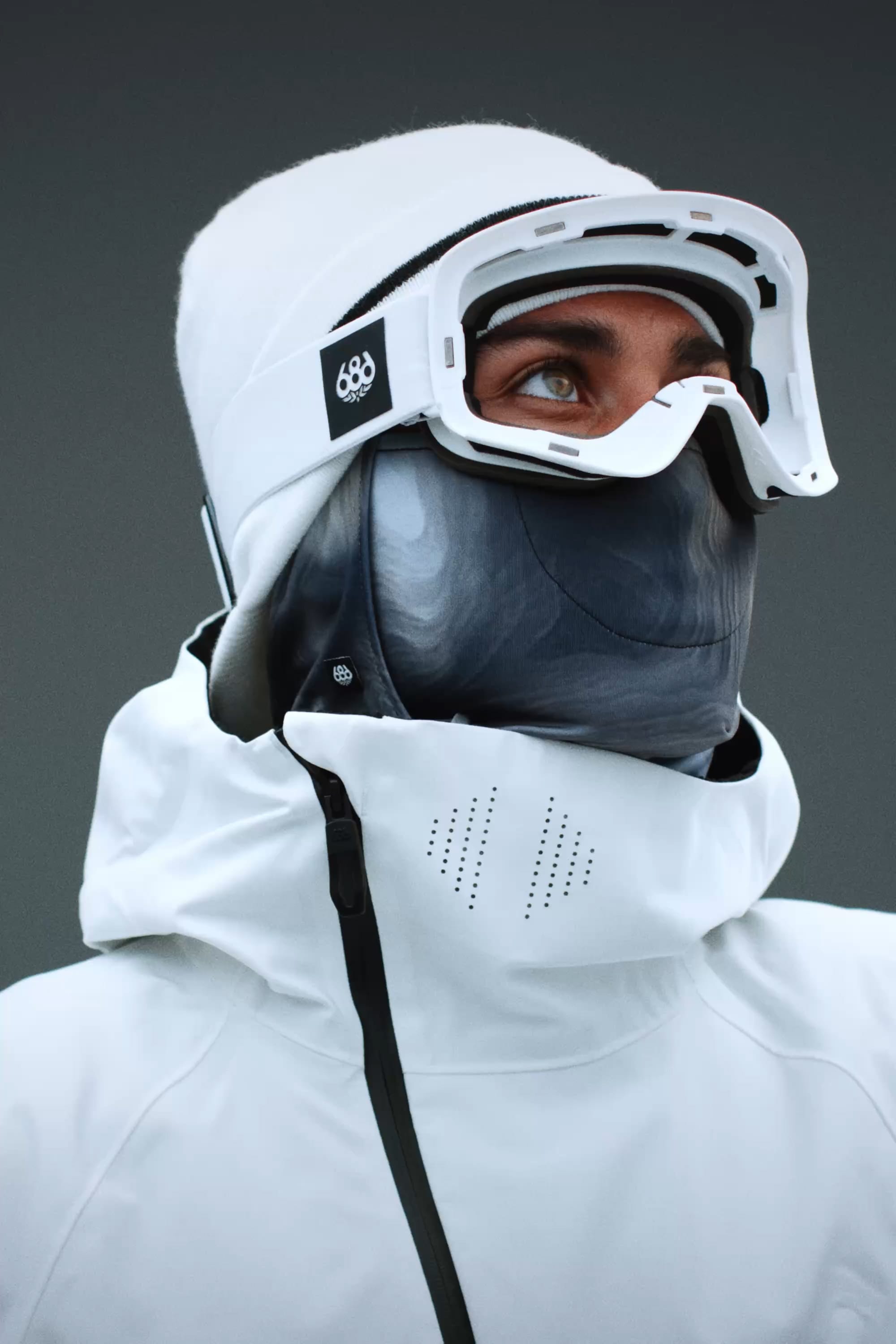 Wearing the 686 SMARTY® Magnetic Goggle by 686, a person in a white jacket and face mask looks upward against a plain gray background, highlighting their winter sport attire.