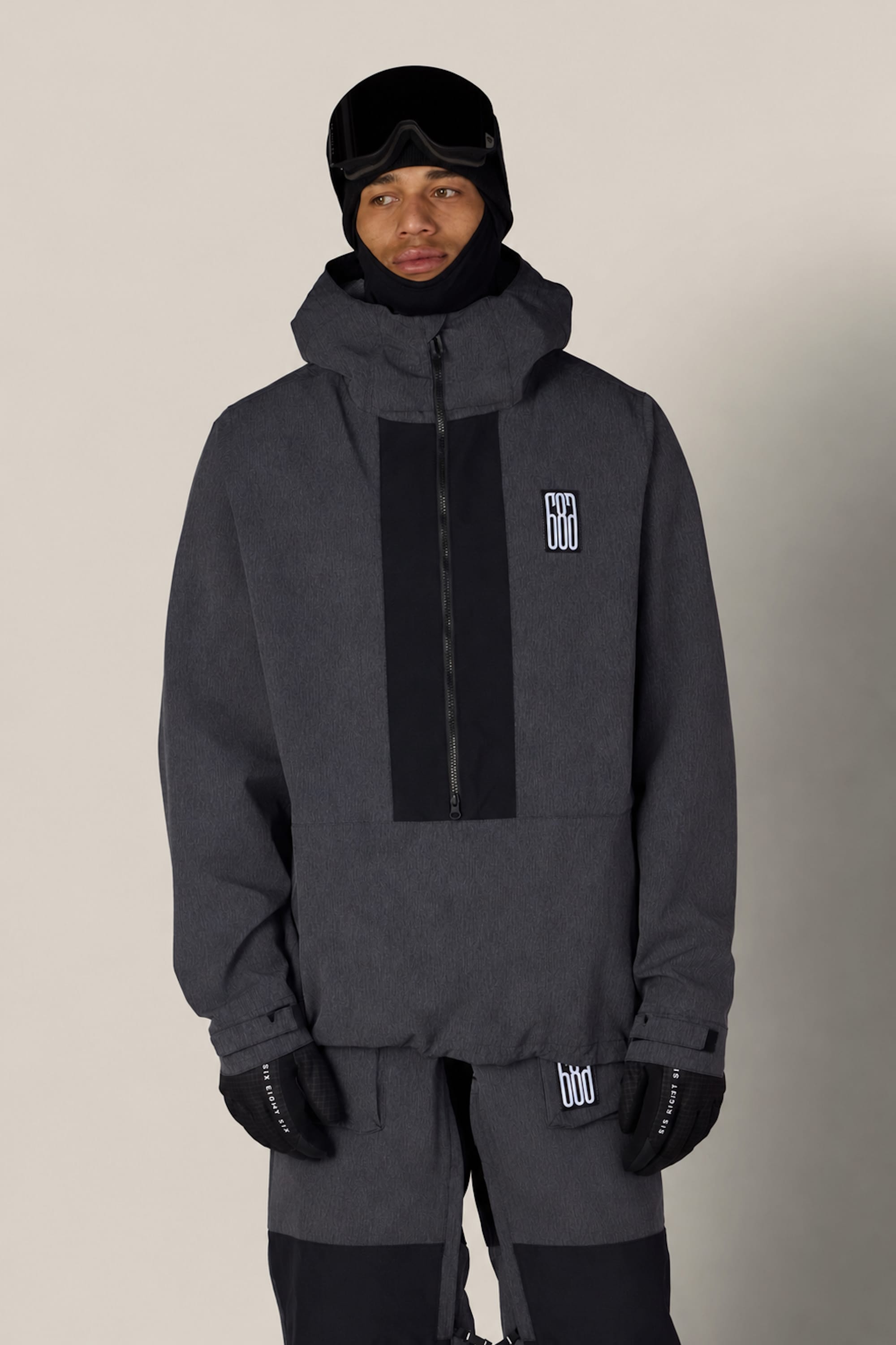 A person wearing the 686 Men’s Ghost™ 2.5L Anorak in dark gray, paired with black gloves, black ski goggles, and a black hat, stands against a plain light background. Both the jacket and pants feature white 686 logos.